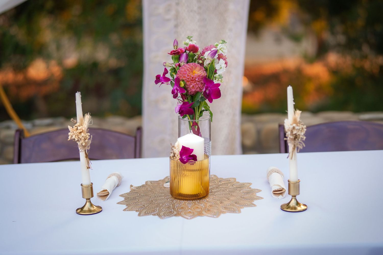 Wedding ceremony table with white linen, gold candlesticks, a floral centerpiece in an amber vase, and a woven doily.