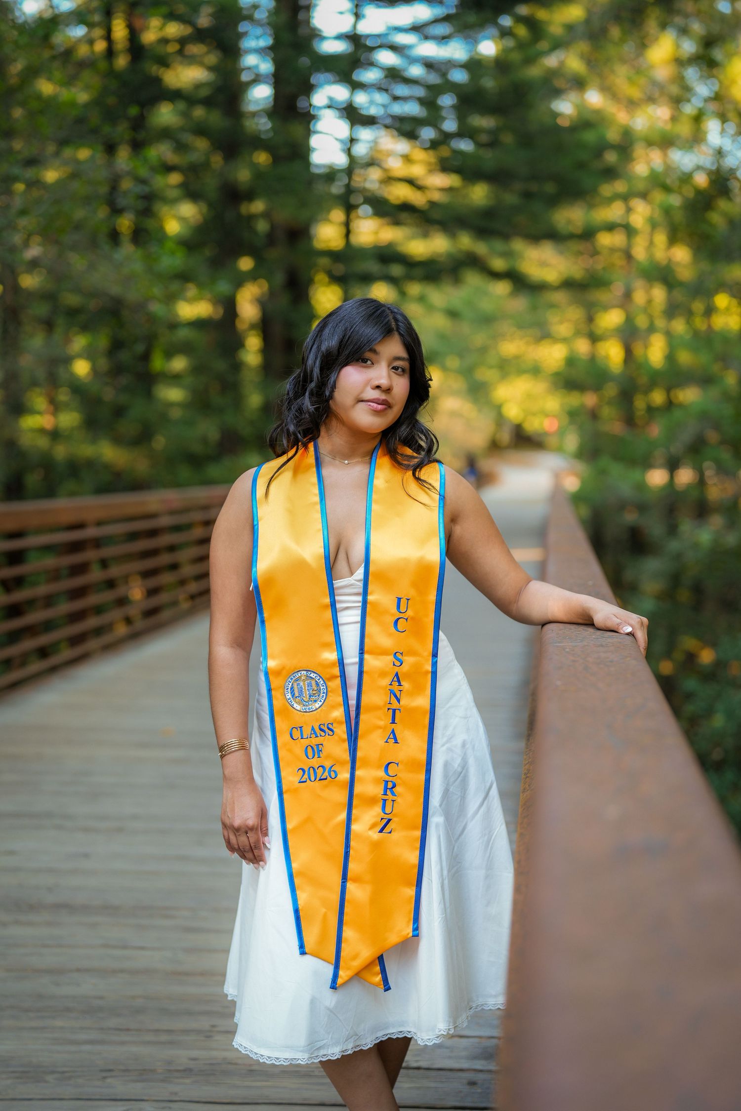 Graduate in a white dress and blue-trimmed gold UC Santa Cruz Class of 2026 stole leans on a bridge railing in the forest.