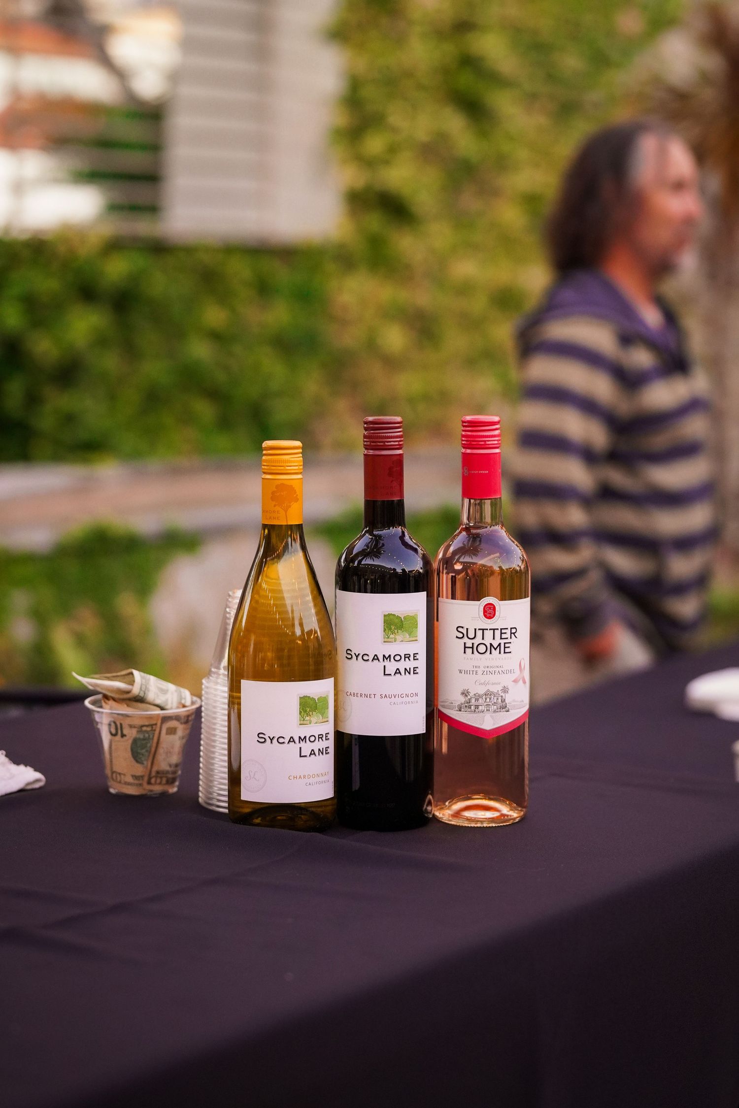 Three bottles of Sycamore Lane and Sutter Home wine stand on a black-draped table beside a tip cup with cash at an outdoor event.
