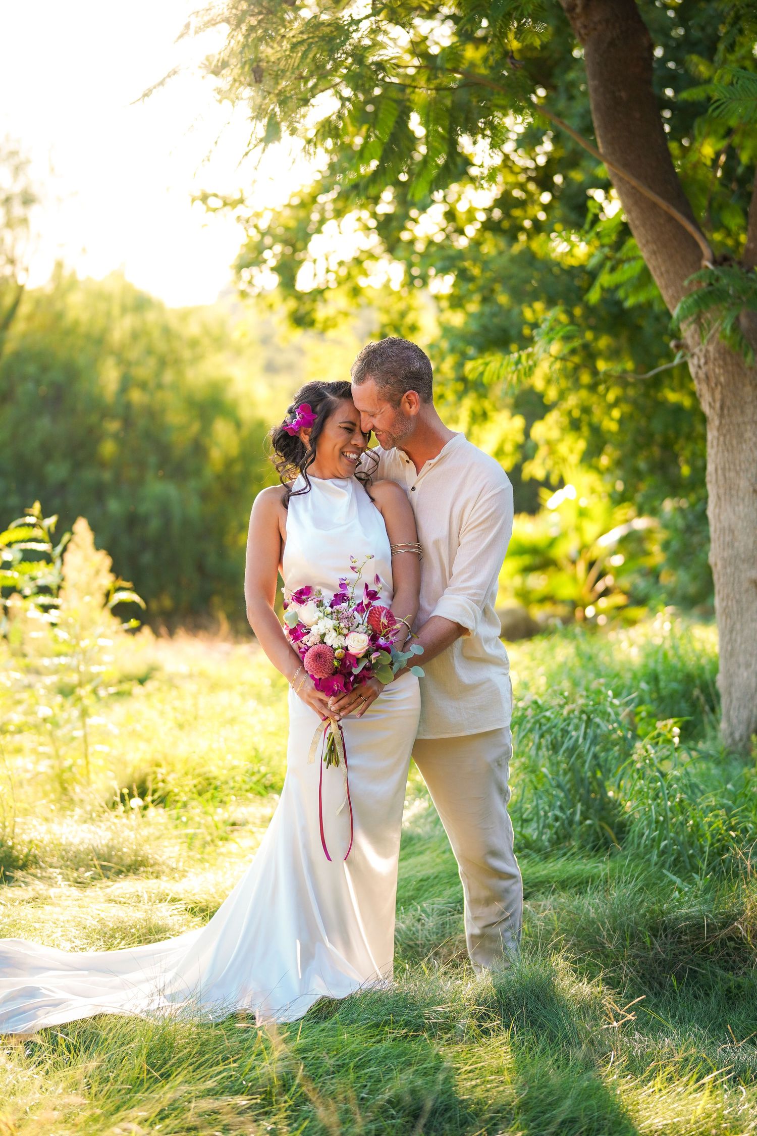 Bride and groom embrace tenderly in golden sunlight beneath a tree, the bride holding a colorful pink and purple bouquet.