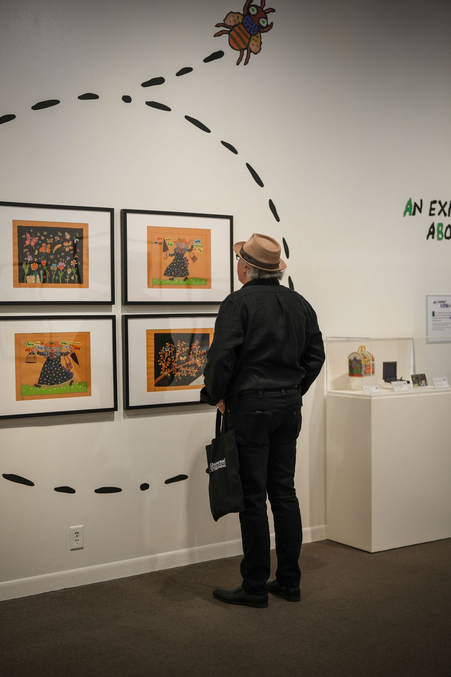 Visitor in a brown hat and black jacket views framed folk-art illustrations on a gallery wall with a dotted bee flight path.
