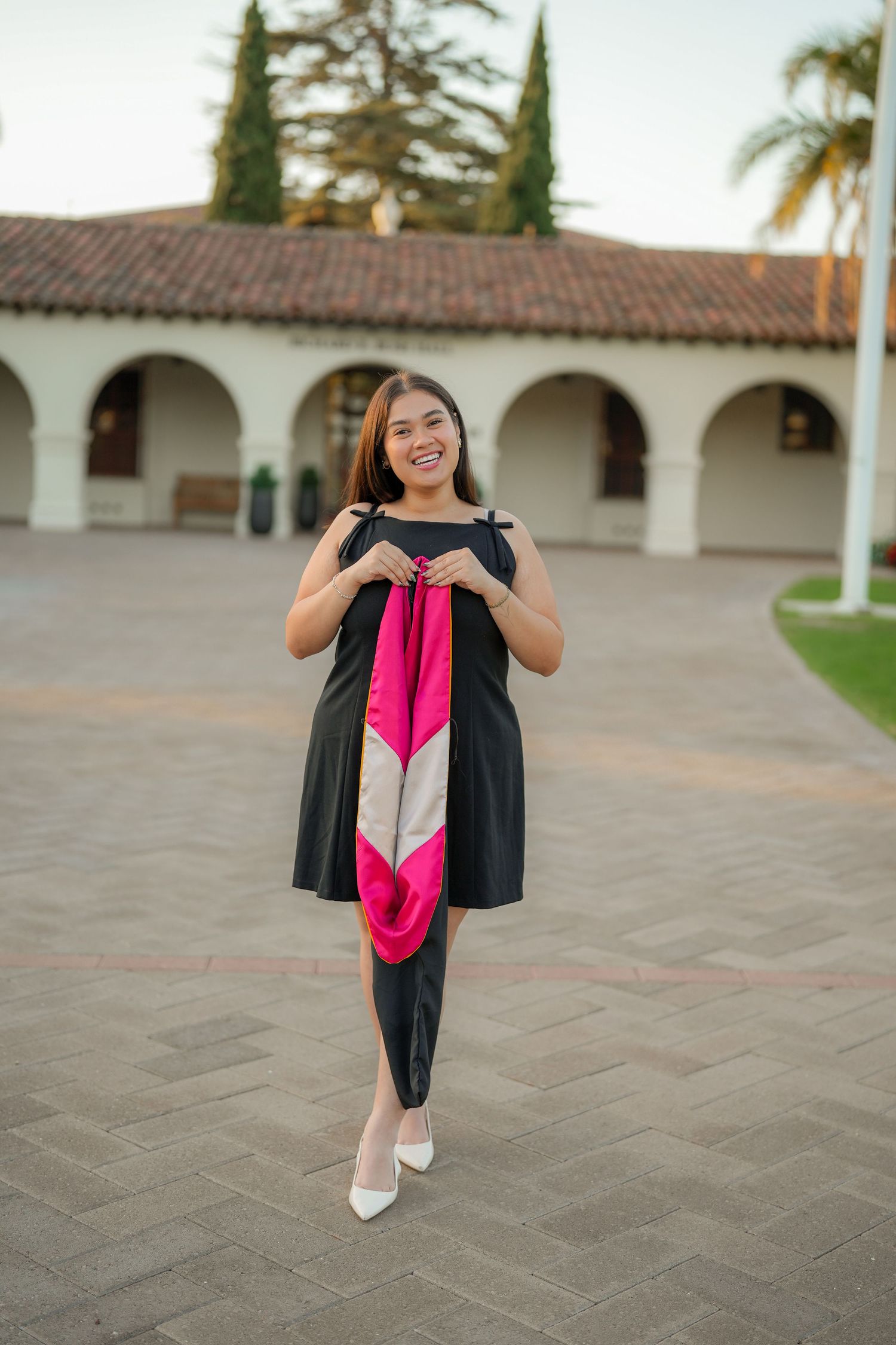 A smiling graduate in a black dress holds a pink and white graduation stole in front of a courtyard with arched, red-roofed buildings.