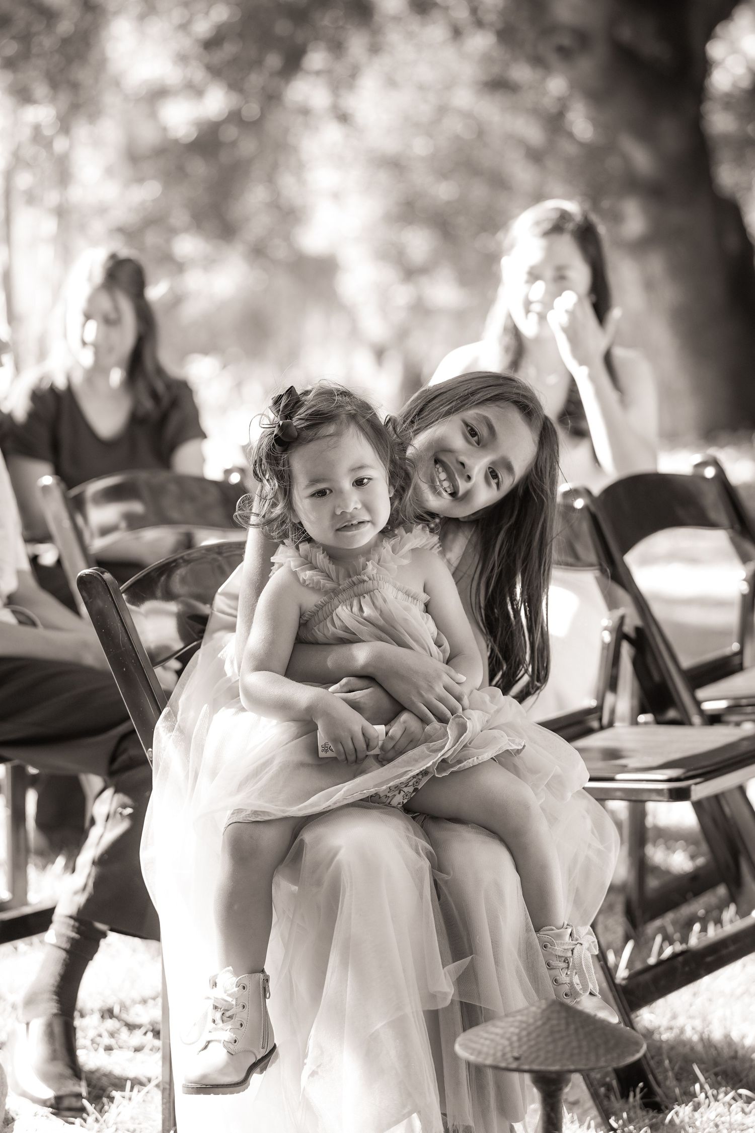A smiling girl in a tulle dress holds a toddler on her lap during an outdoor wedding ceremony in this black and white photo.