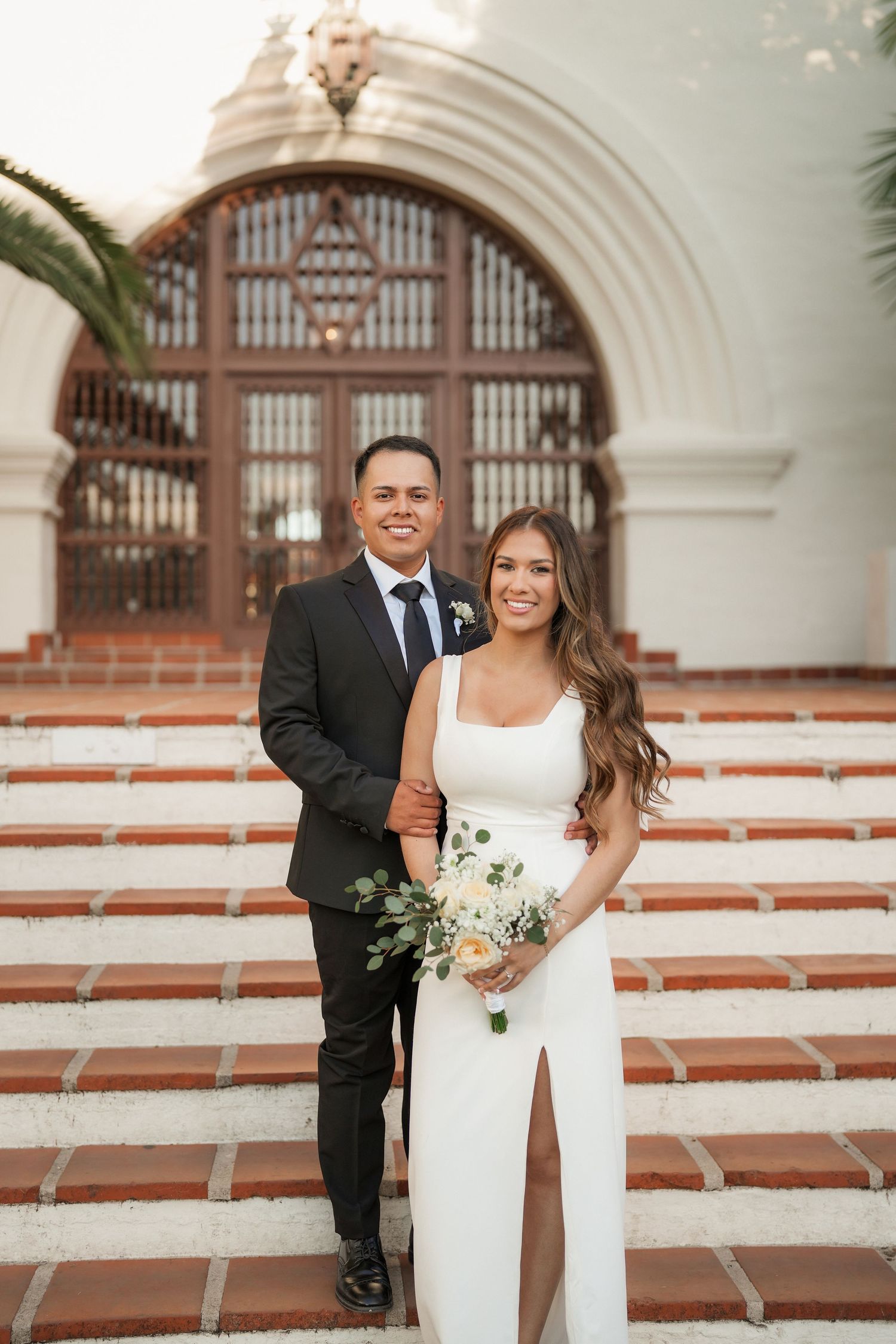 Smiling bride with a bouquet and groom in a dark suit stand close on red-tiled steps before an ornate arched wooden door.
