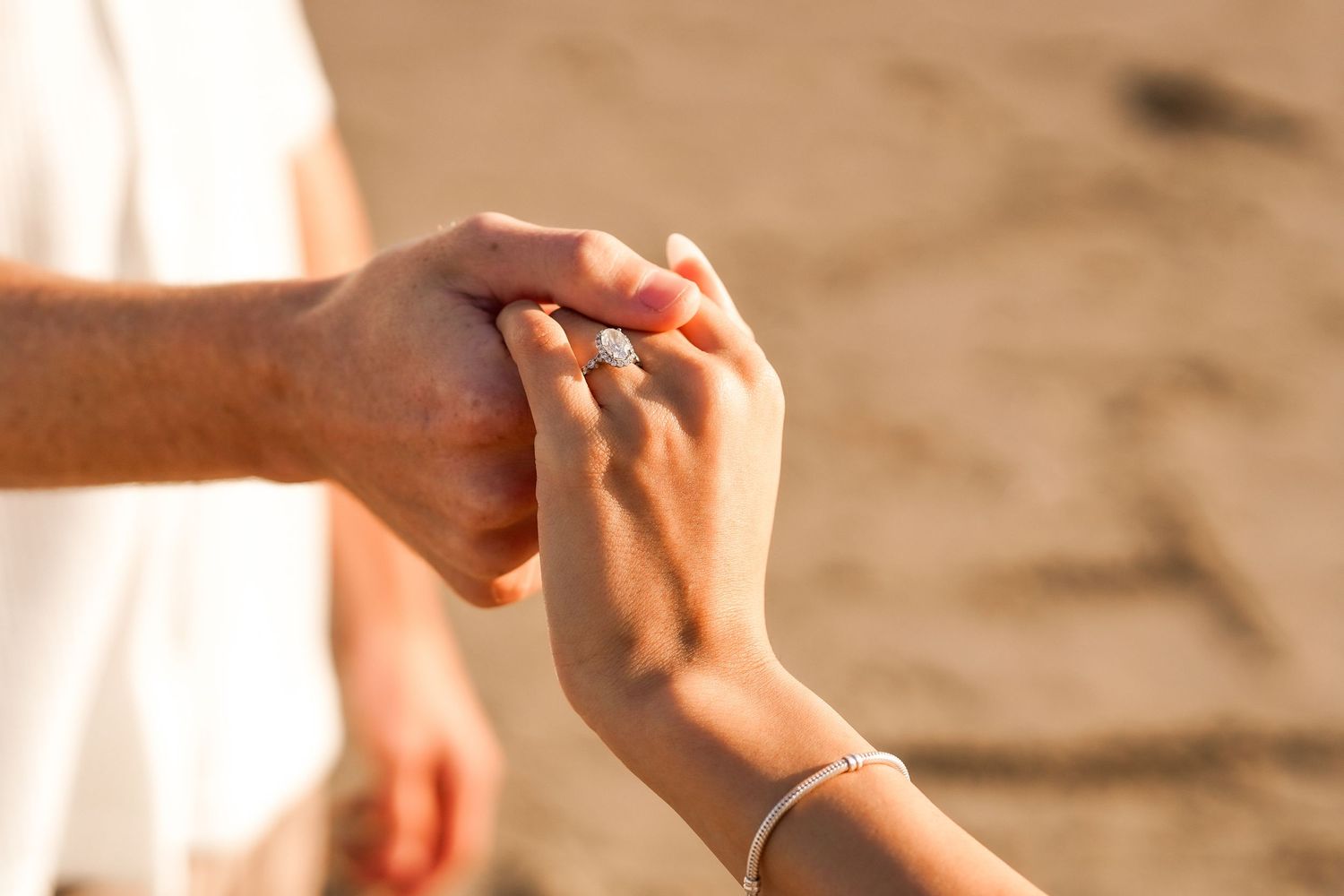 Close-up of a couple holding hands as a diamond engagement ring sparkles on the woman's finger on a sandy beach.