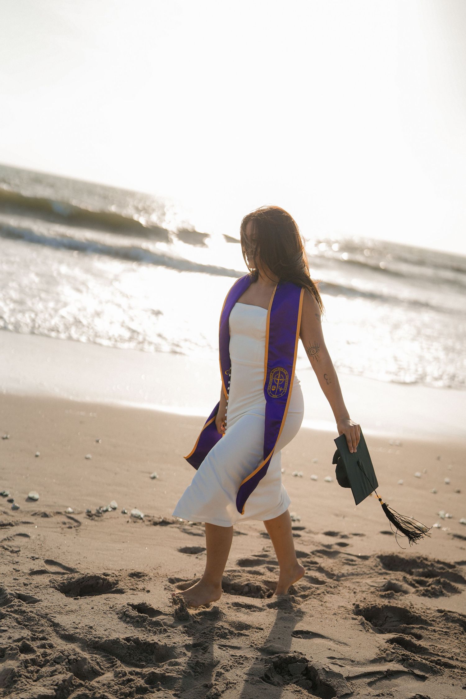 Barefoot graduate in a white dress and purple stole walks along the sandy beach holding her green graduation cap.