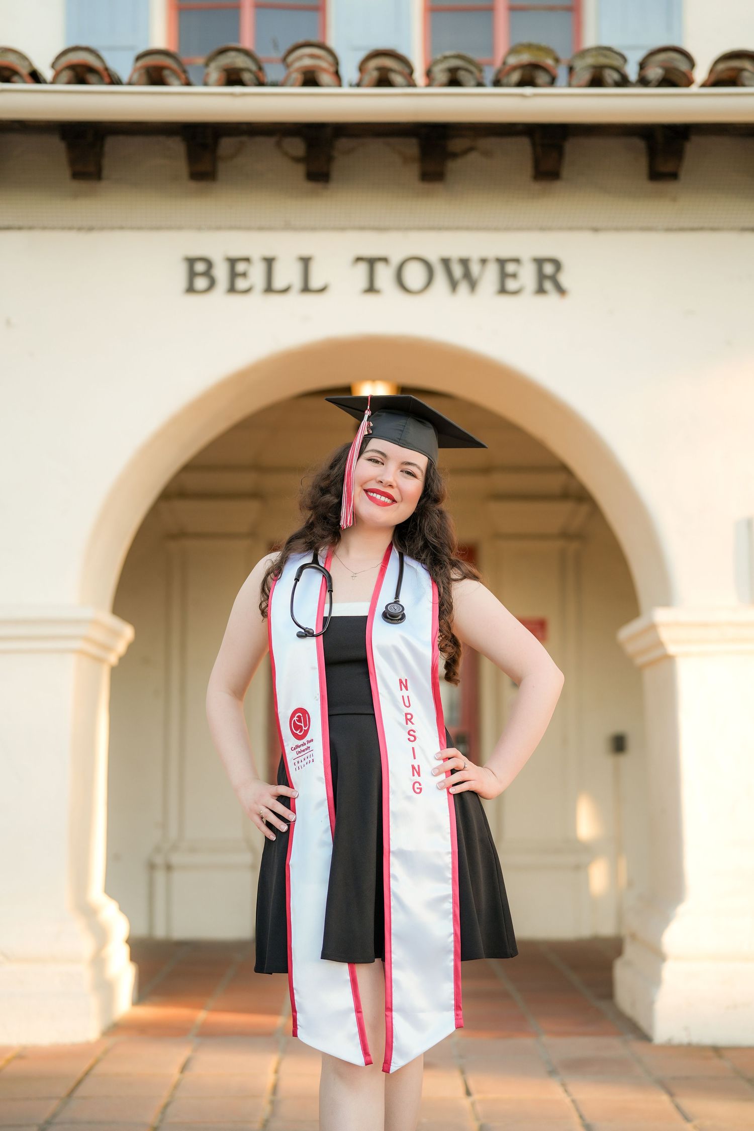 Nursing graduate in cap, sash and stethoscope poses with hands on hips beneath the Bell Tower archway on a sunny campus.
