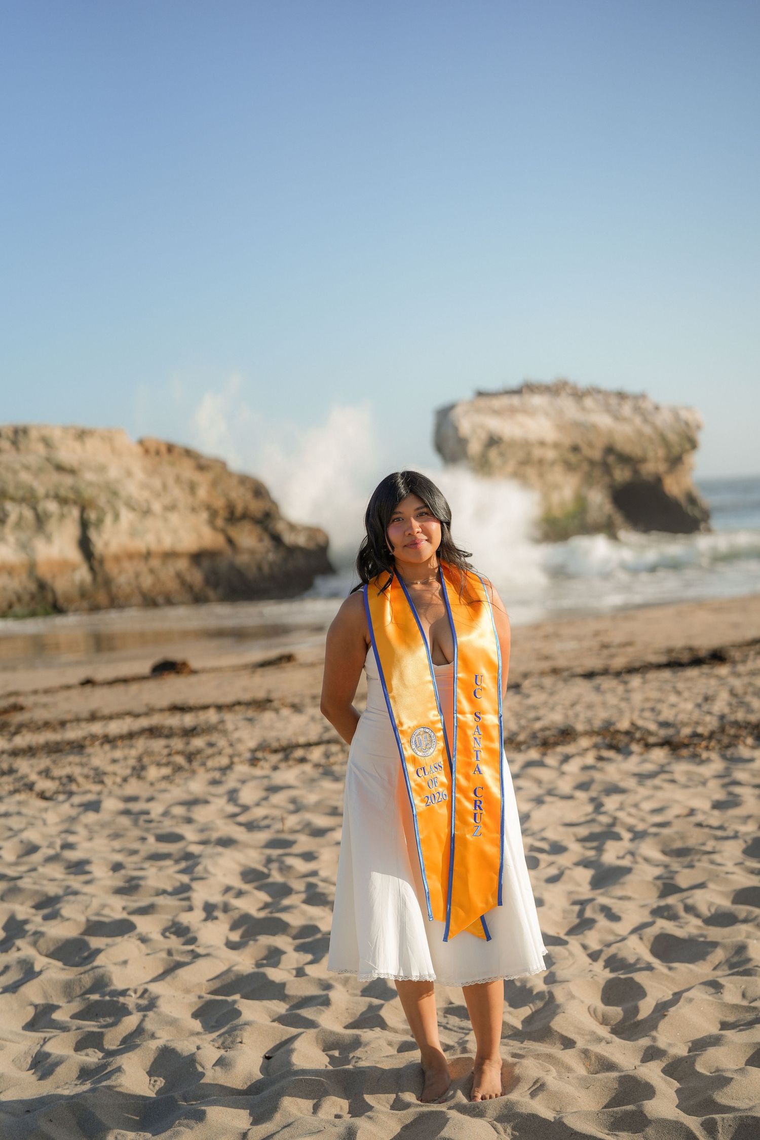 Barefoot graduate in a white dress and orange stole stands on a sandy beach with rock formations and ocean waves behind.
