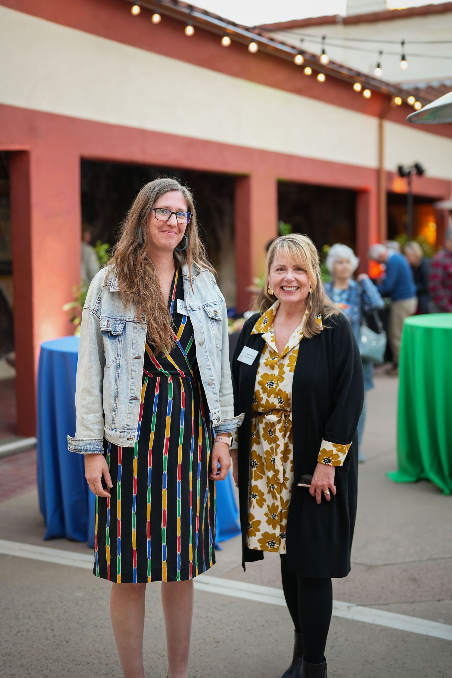 Two smiling women wearing name tags pose together at an outdoor evening event with string lights and draped tables.