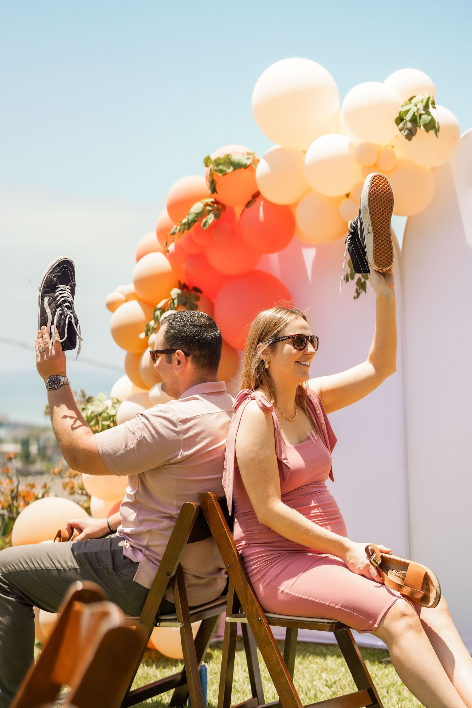Smiling pregnant woman in a pink dress and a man in sunglasses raise shoes in the air at an outdoor party with peach balloon garland.