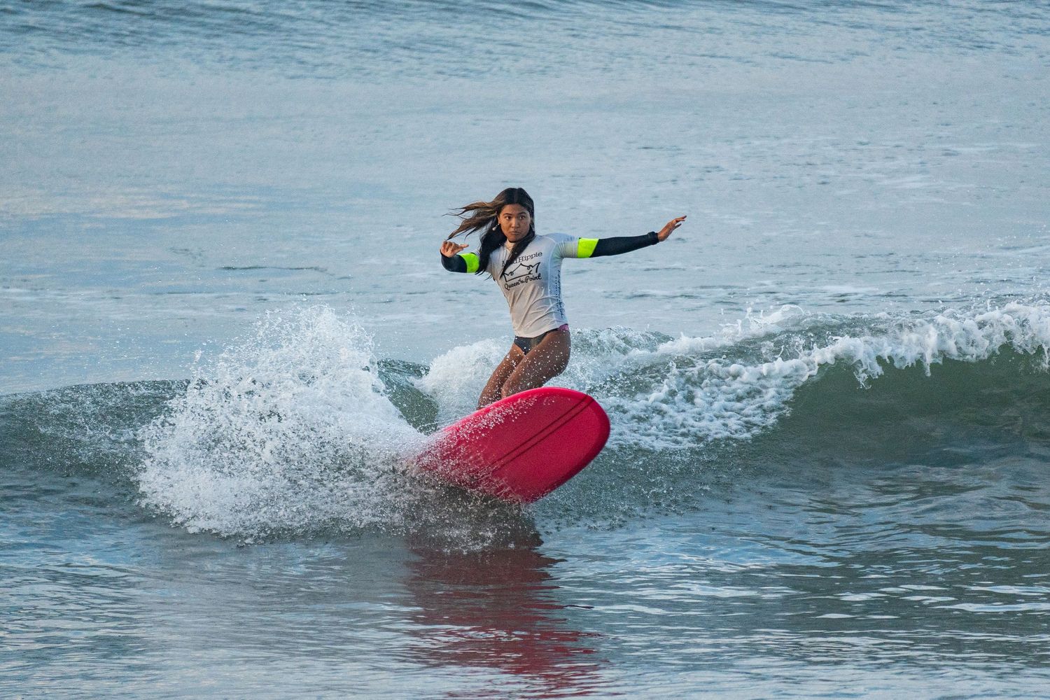Female surfer in a gray top carves a turn on a red surfboard, sending spray off a breaking wave.