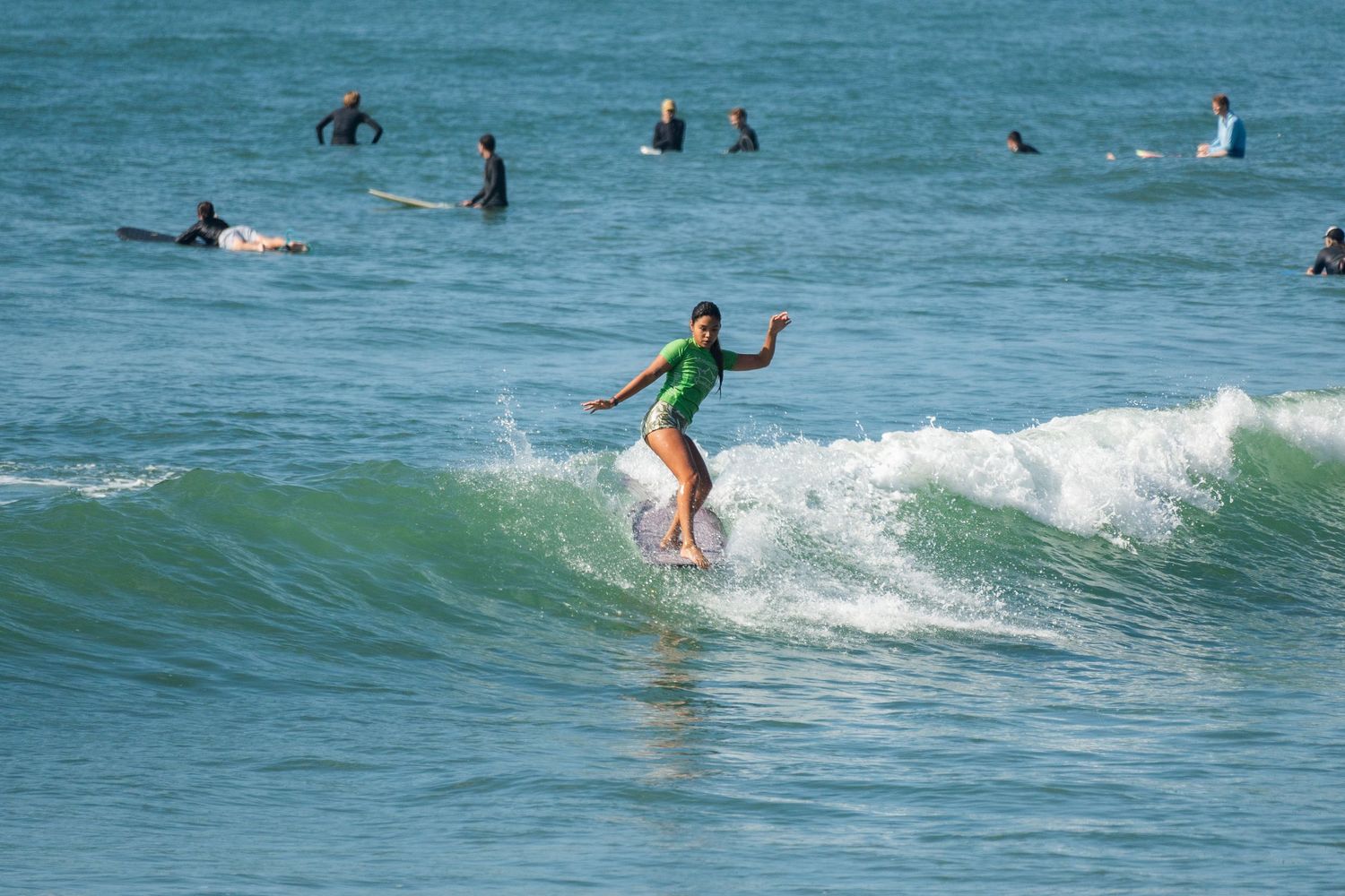 A surfer in a green top rides a longboard down a wave while many surfers wait in the lineup behind.