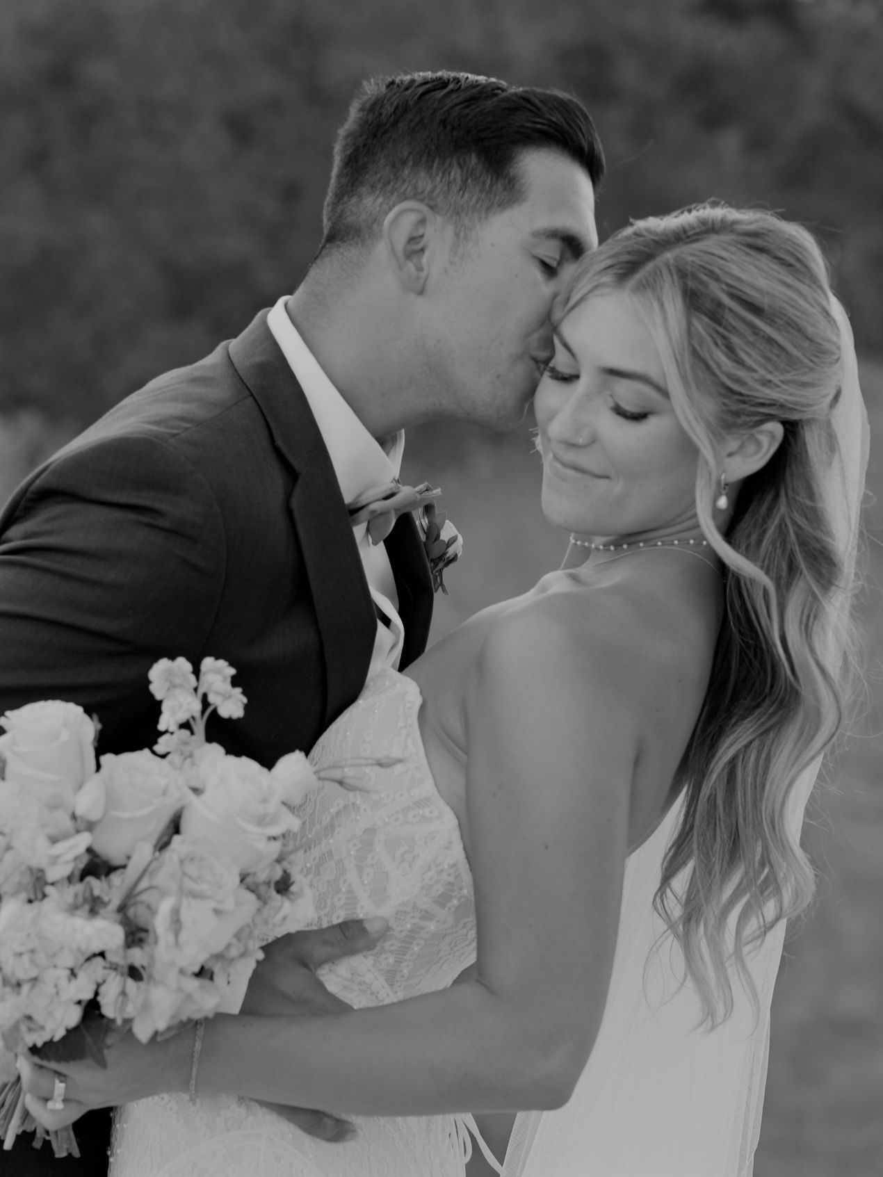 Black and white wedding portrait of a groom kissing his bride's temple as she smiles and holds a bouquet of white flowers.