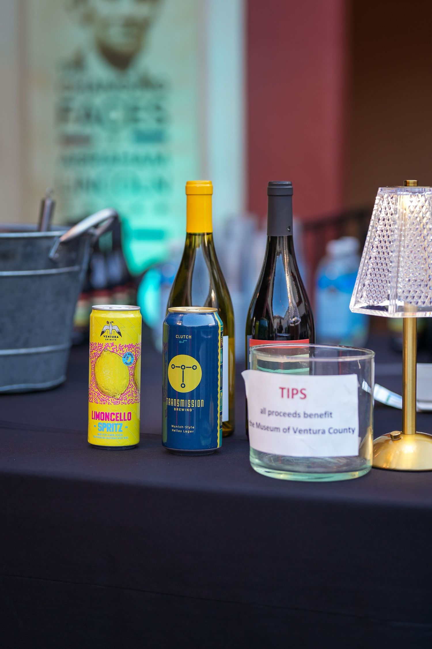 Bar table with canned limoncello spritz, wine bottles, a wine bucket and a tip jar noting proceeds benefit the Museum of Ventura County.