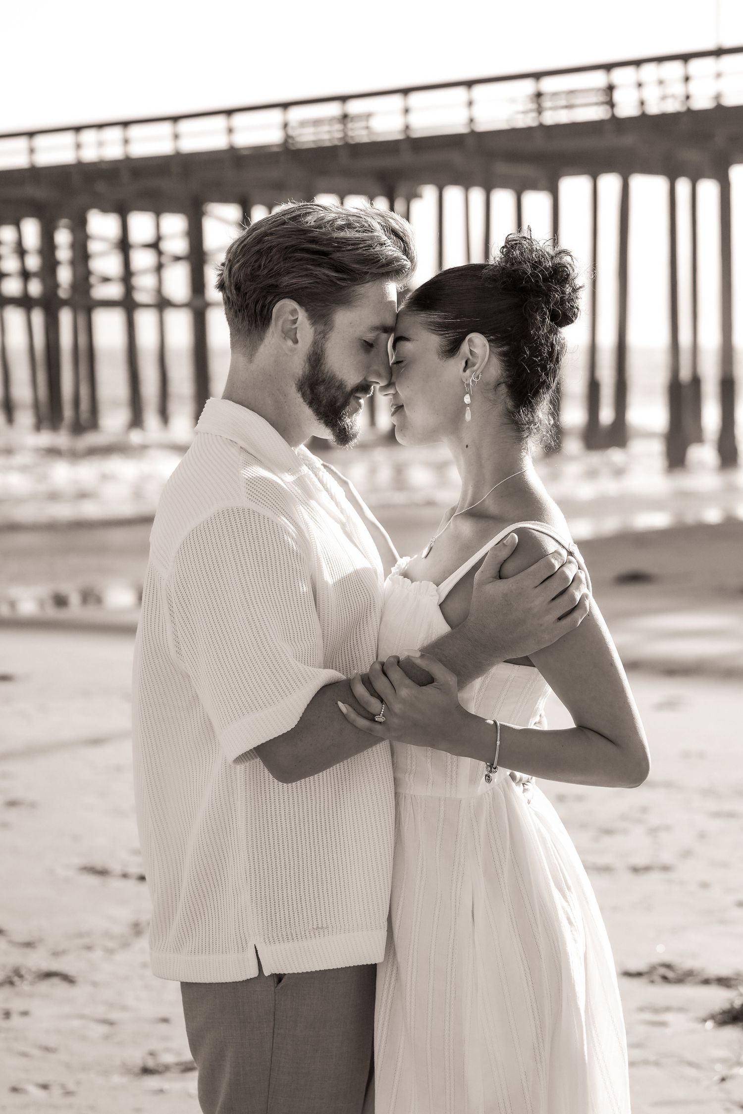 Sepia engagement photo of a couple embracing forehead to forehead on the beach in front of a wooden pier.