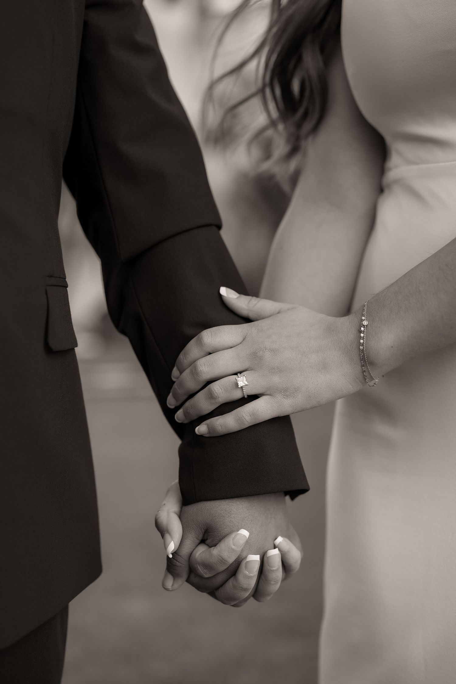 Black and white close-up of a bride's hand with a diamond engagement ring resting on the groom's sleeve as they hold hands.