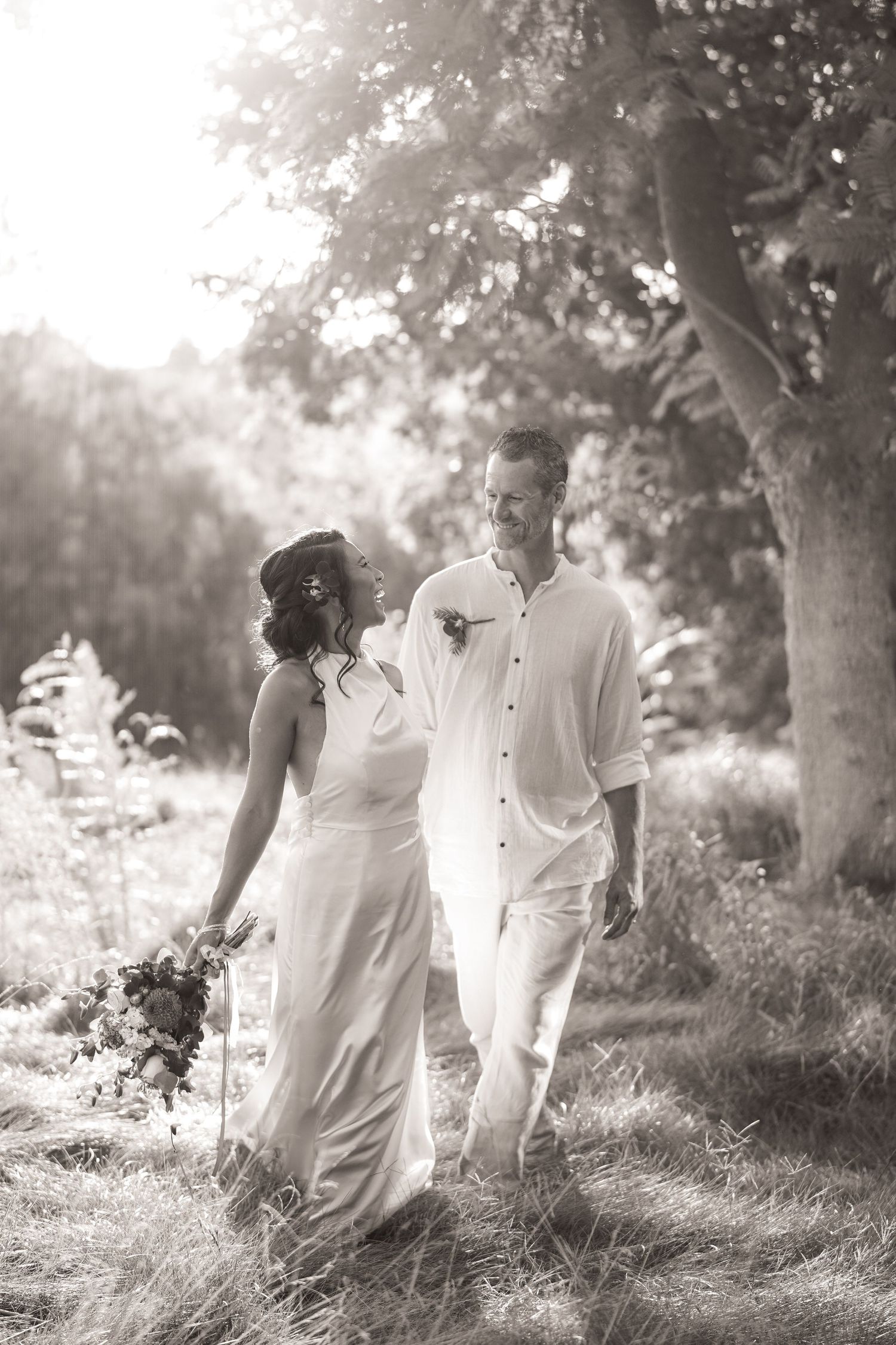 Black and white image of a bride and groom walking hand in hand through tall grass beneath a large shady tree.