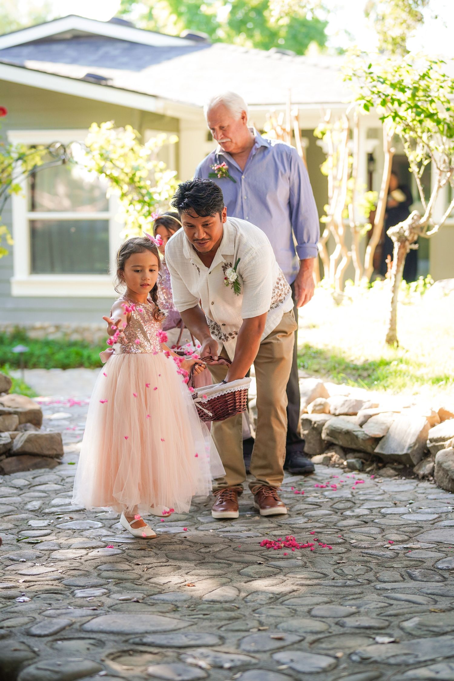 A flower girl in a blush tulle dress scatters petals from a basket along a stone path with a man guiding her at a garden wedding.