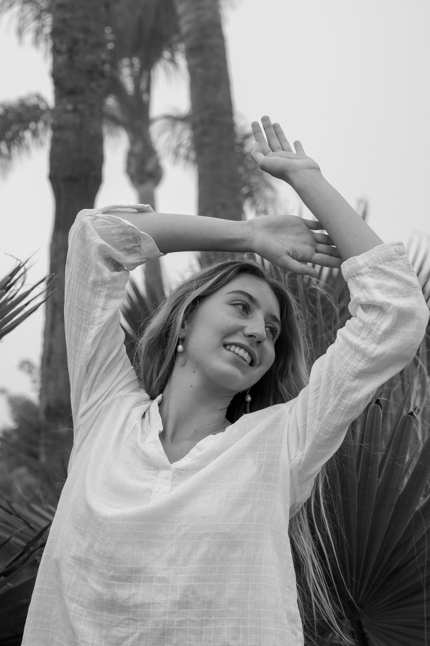 Black and white image of a smiling woman in a white tunic raising her arms among palm trees and fronds.