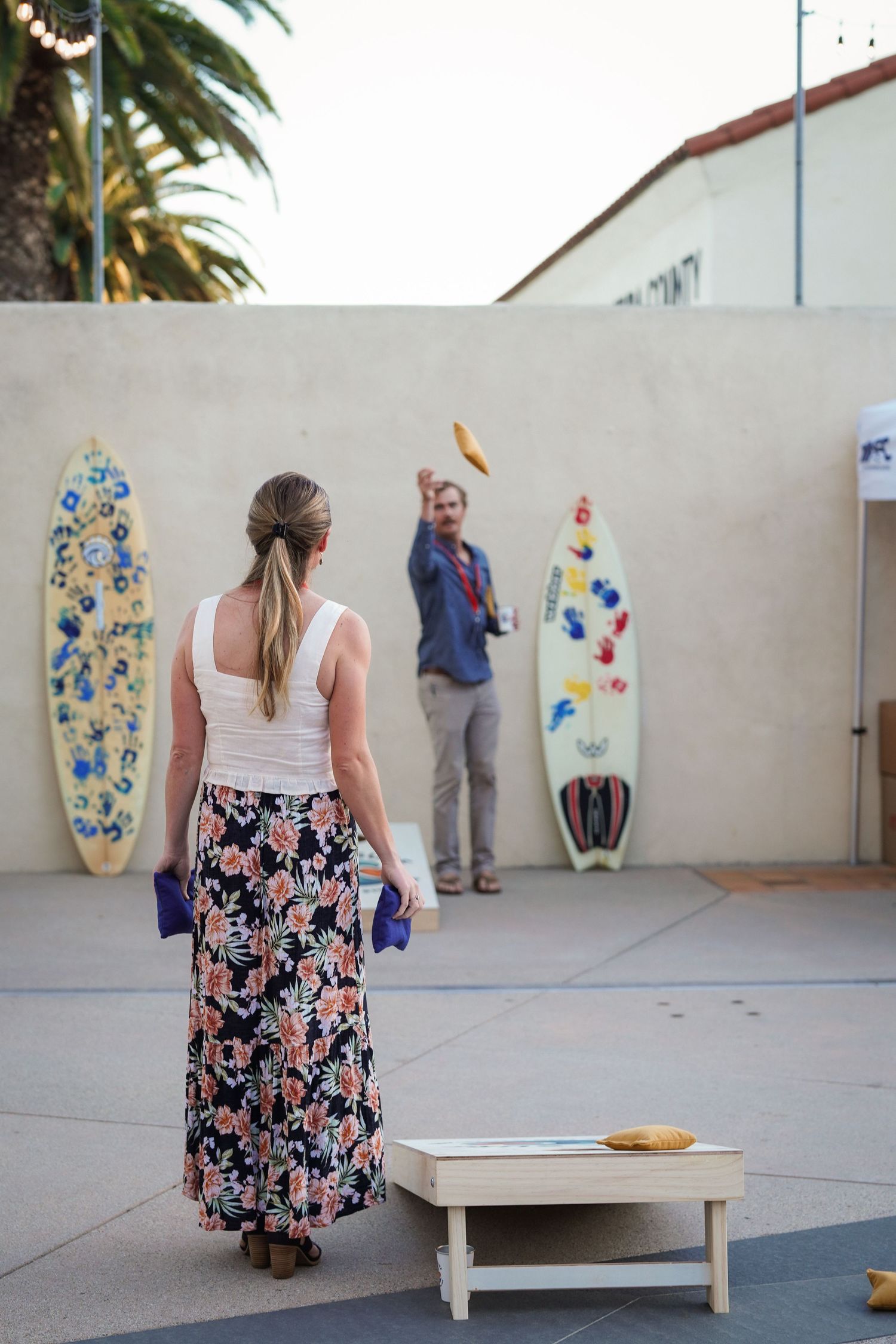 A woman in a floral skirt watches a man toss a cornhole bag near painted surfboards at an outdoor beach-themed event.