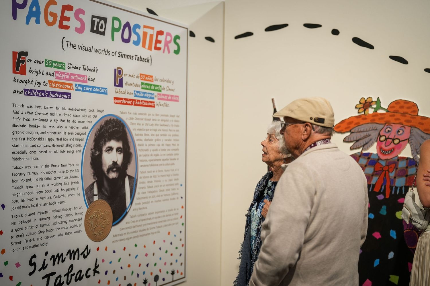 Two visitors study a Pages to Posters exhibit panel about the visual work of Simms Taback beside a colorful illustration.