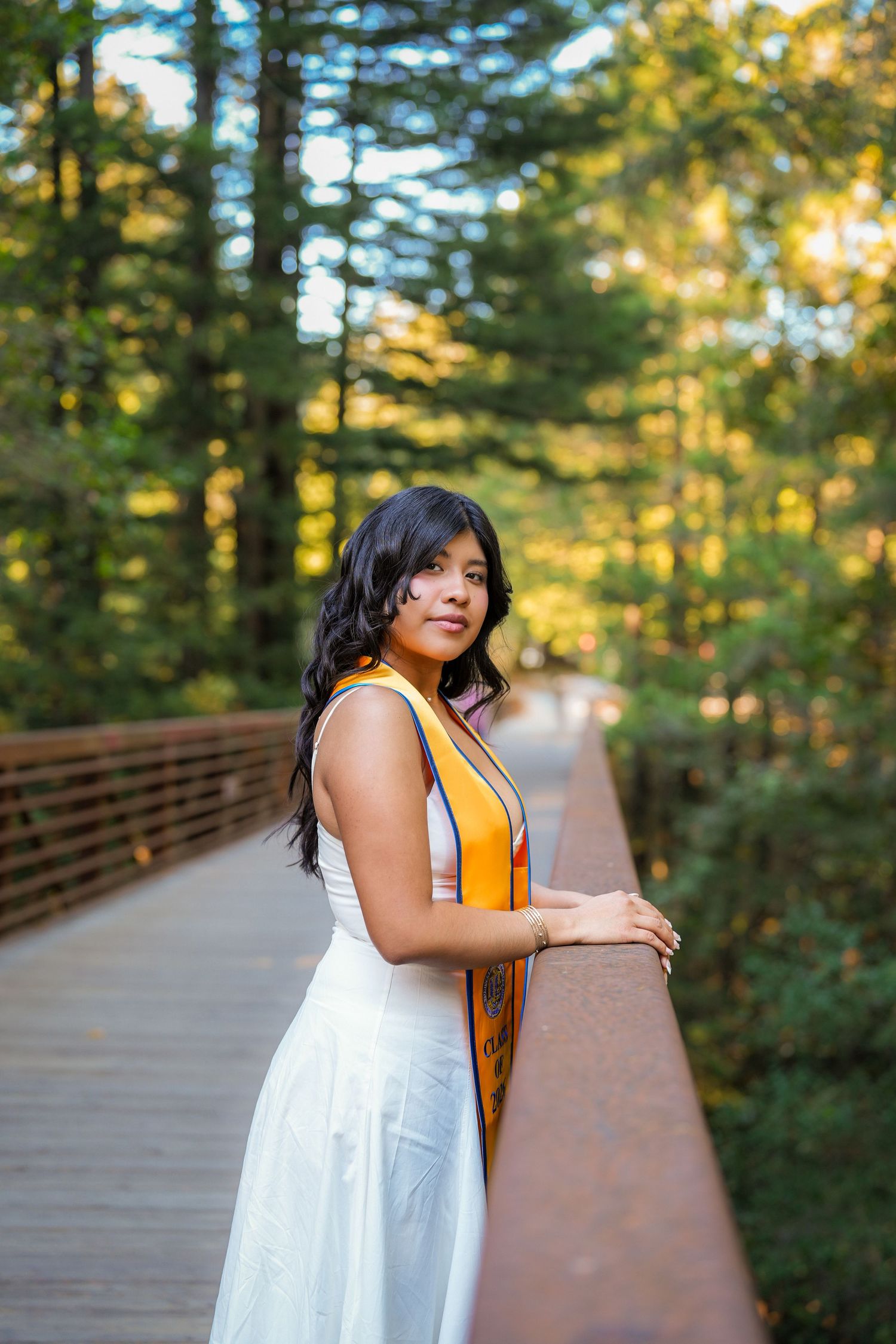 Graduate in a white dress and orange stole leans on a bridge railing in a golden-lit forest.