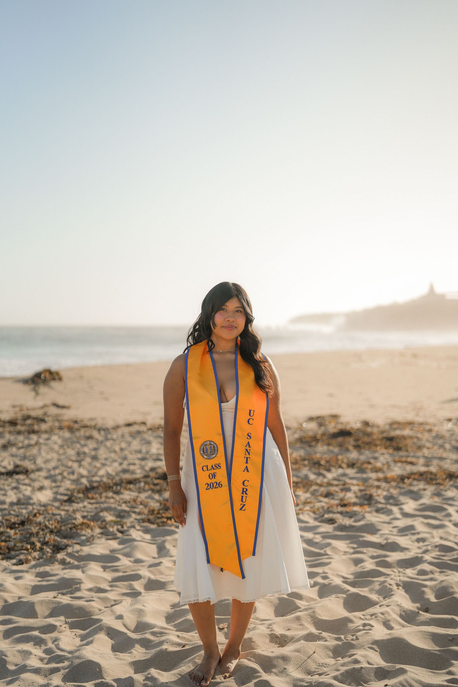Graduate in a white dress wearing an orange UC Santa Cruz Class of 2026 sash while standing on a sandy beach.