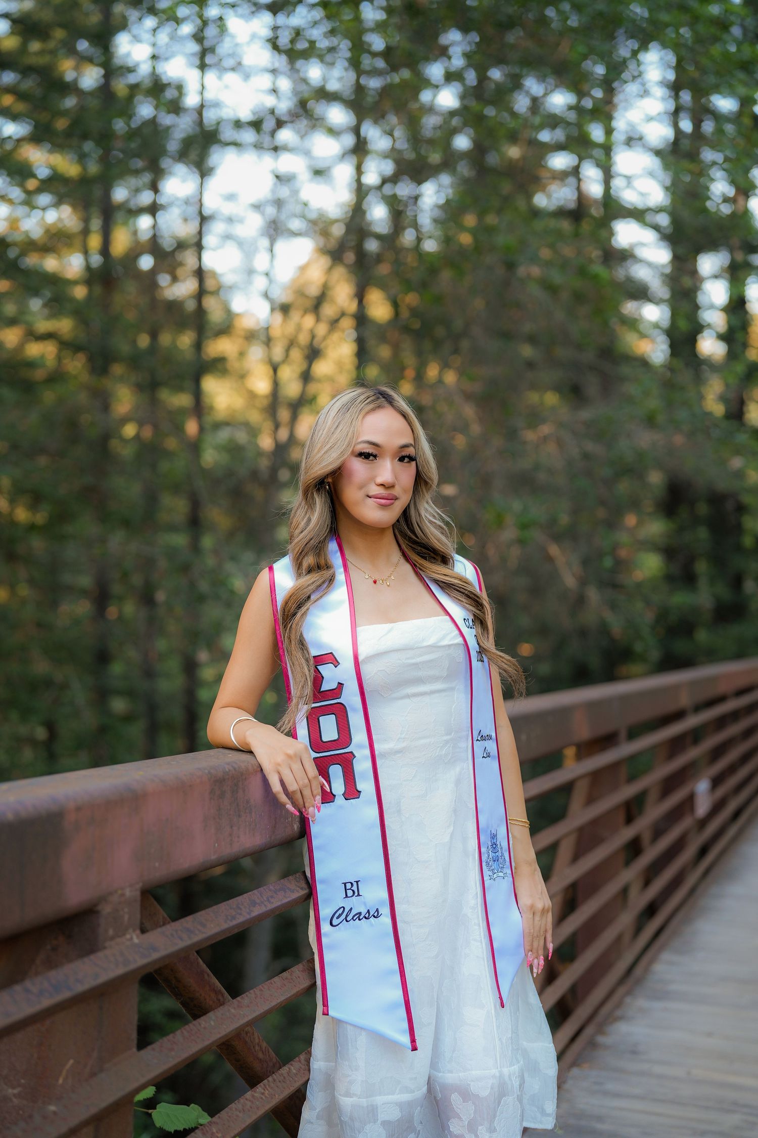 Graduate in a white dress and white and maroon class stole smiles while leaning on a wooden bridge railing in a forest.