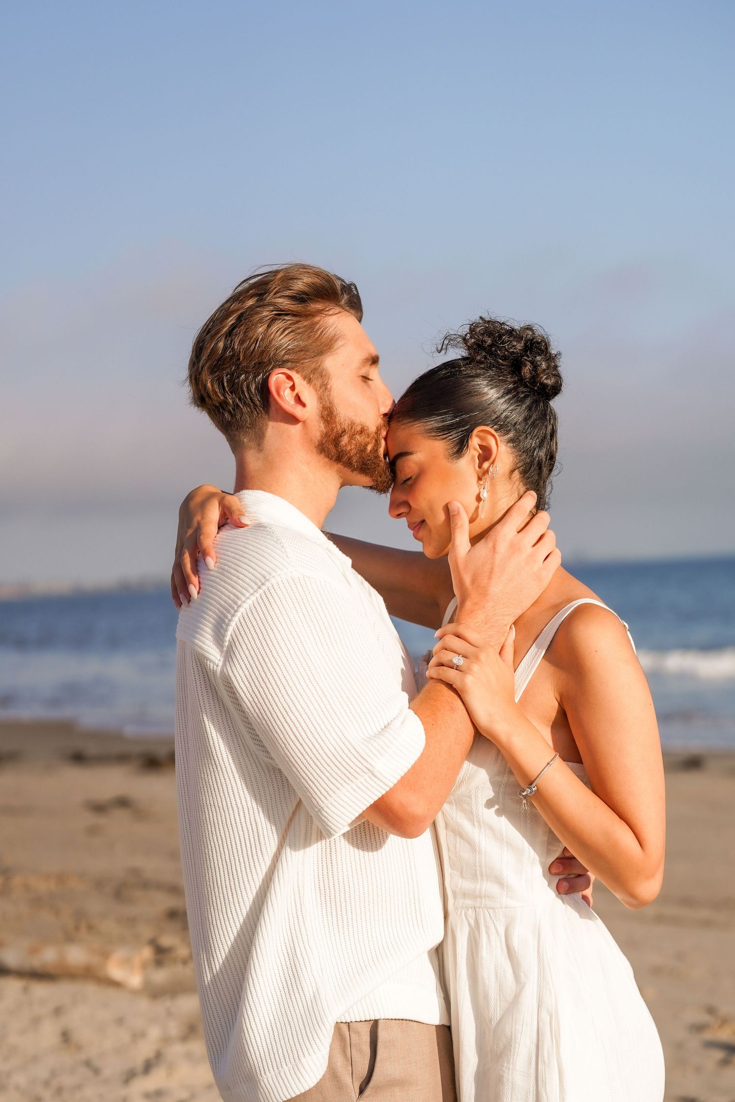 Engaged couple in white outfits embrace on a sunny beach as the man kisses her forehead, with the ocean in the background.