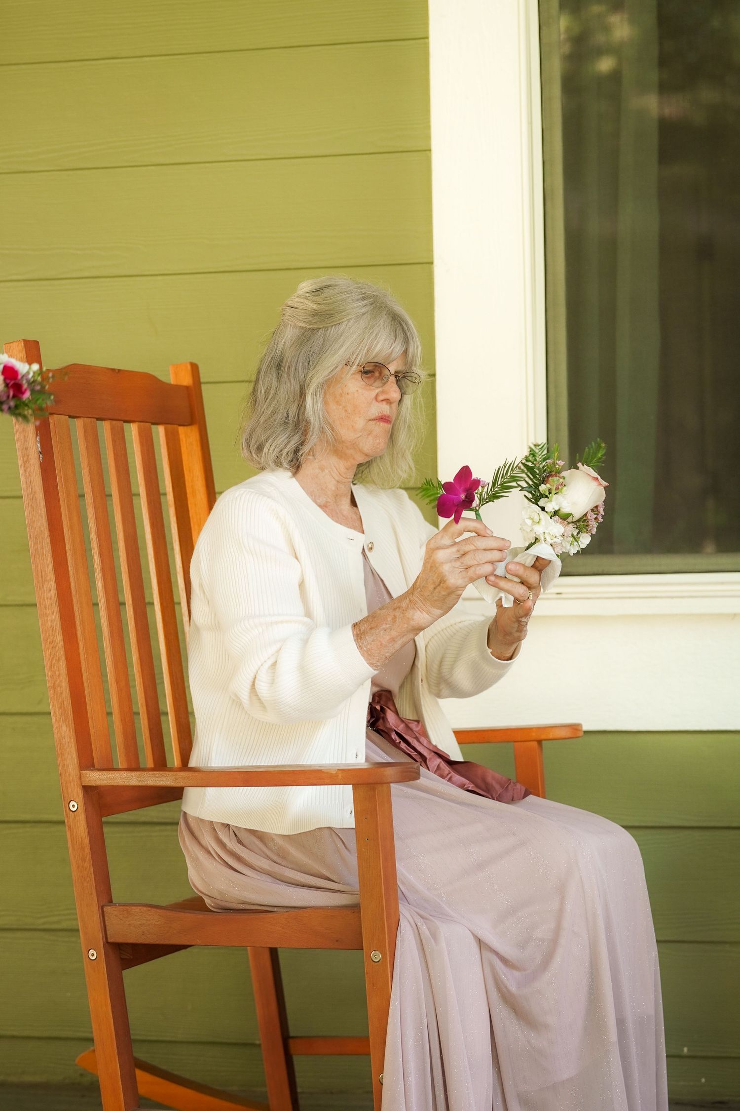 Older woman with gray hair and glasses sits in an orange rocking chair on a porch, admiring a small pink and white bouquet.