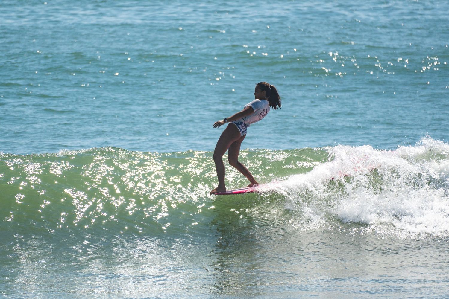 A surfer in a swimsuit leans back on a red longboard while riding a sparkling turquoise wave.
