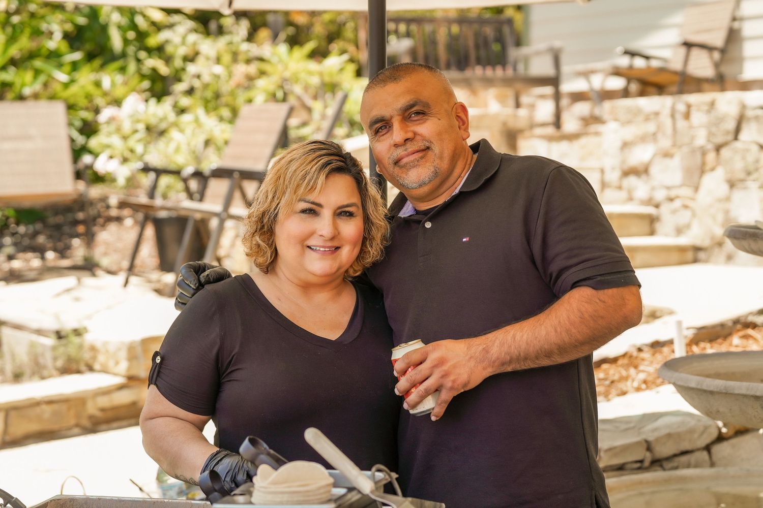 Smiling man and woman in black shirts stand close together at an outdoor patio with stone walls while preparing food at a table.