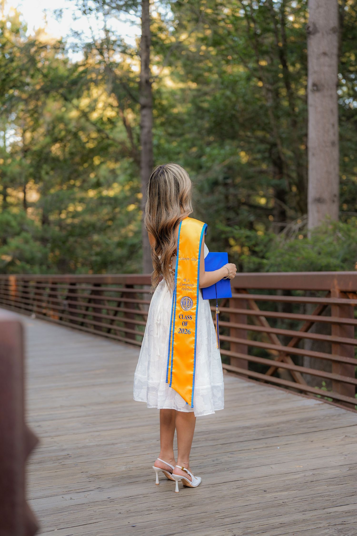 Graduate with long wavy hair and a gold sash faces away on a wooden bridge in a forest, holding a blue graduation cap.