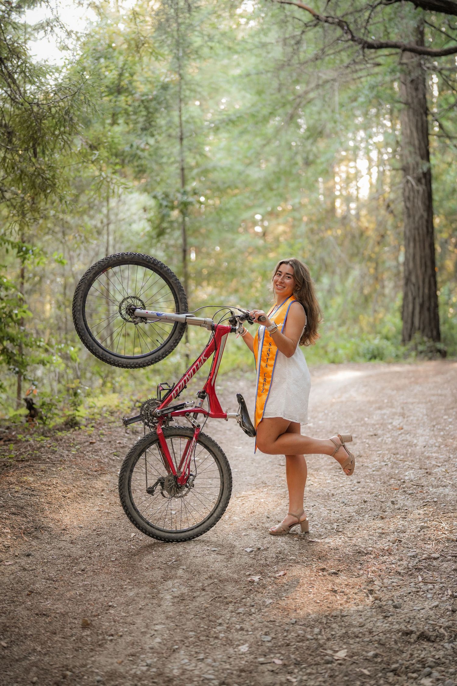 Playful graduate in a gold stole and heels lifts a red mountain bike's front wheel on a sunlit forest trail.