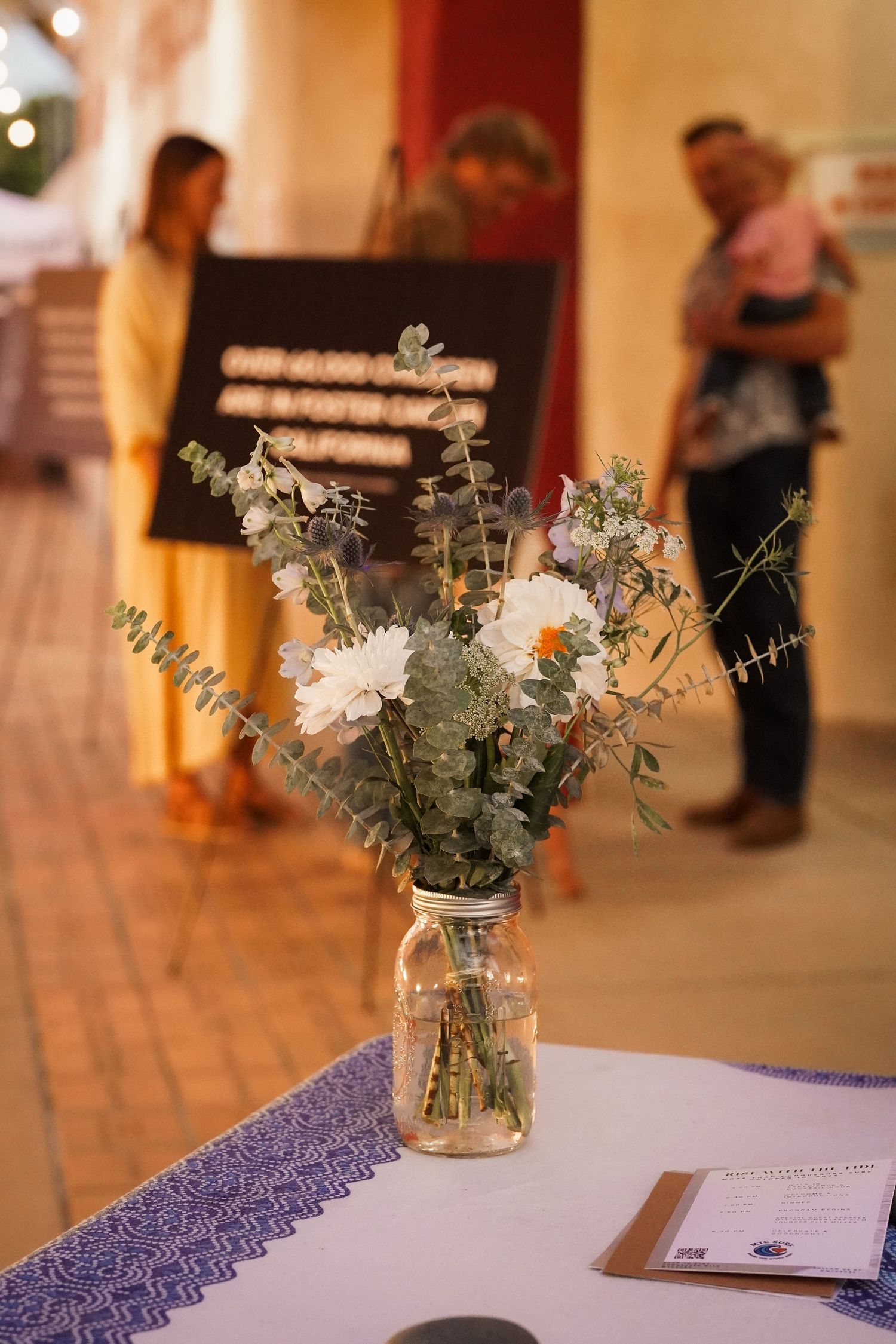 Mason jar vase of eucalyptus and white flowers on a table with guests and a chalkboard sign blurred behind it at an evening event.