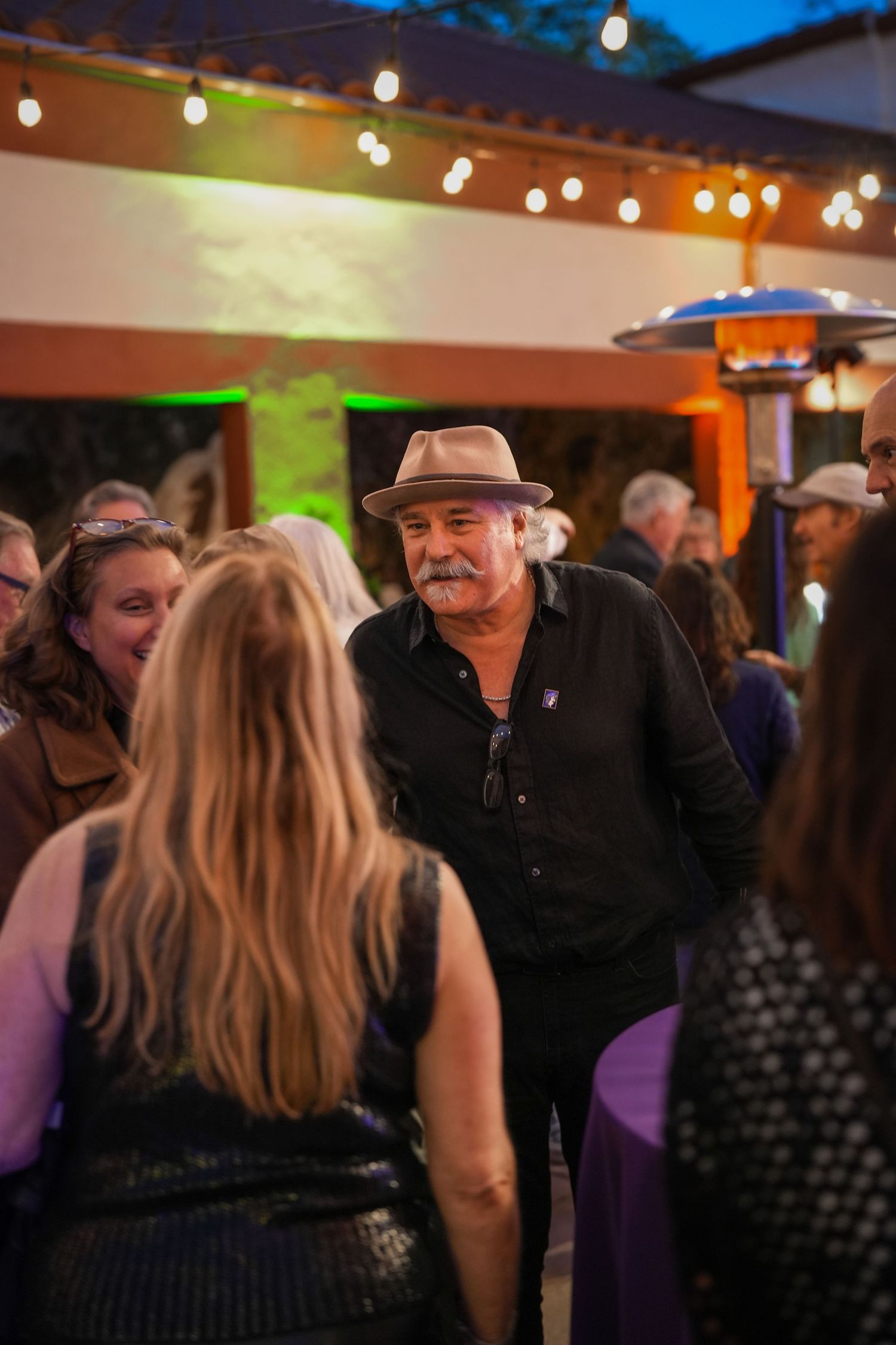 A mustached man in a tan hat chats with guests at a lively outdoor evening reception under warm string lights.