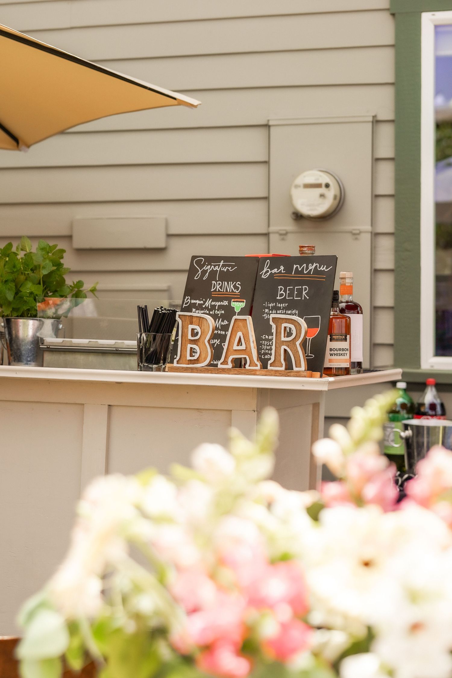 Outdoor wedding bar with wooden BAR letters, chalkboard drink menus, bottles, and pink and white flowers in the foreground.