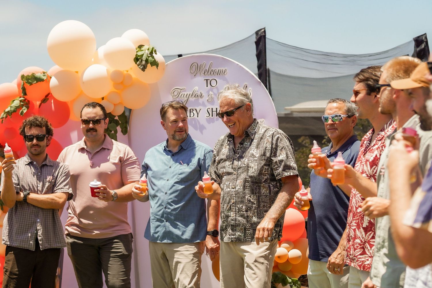 Group of smiling men holding drinks beside an orange and white balloon arch and welcome sign at an outdoor celebration.
