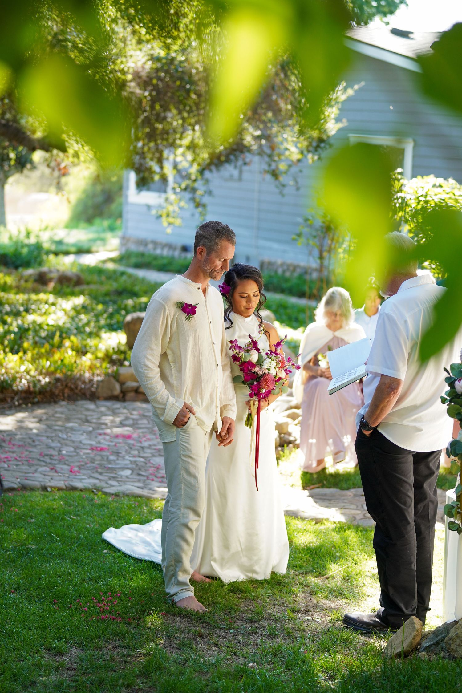 A barefoot bride holding a pink bouquet and a groom in linen stand before an officiant during an intimate garden wedding ceremony.