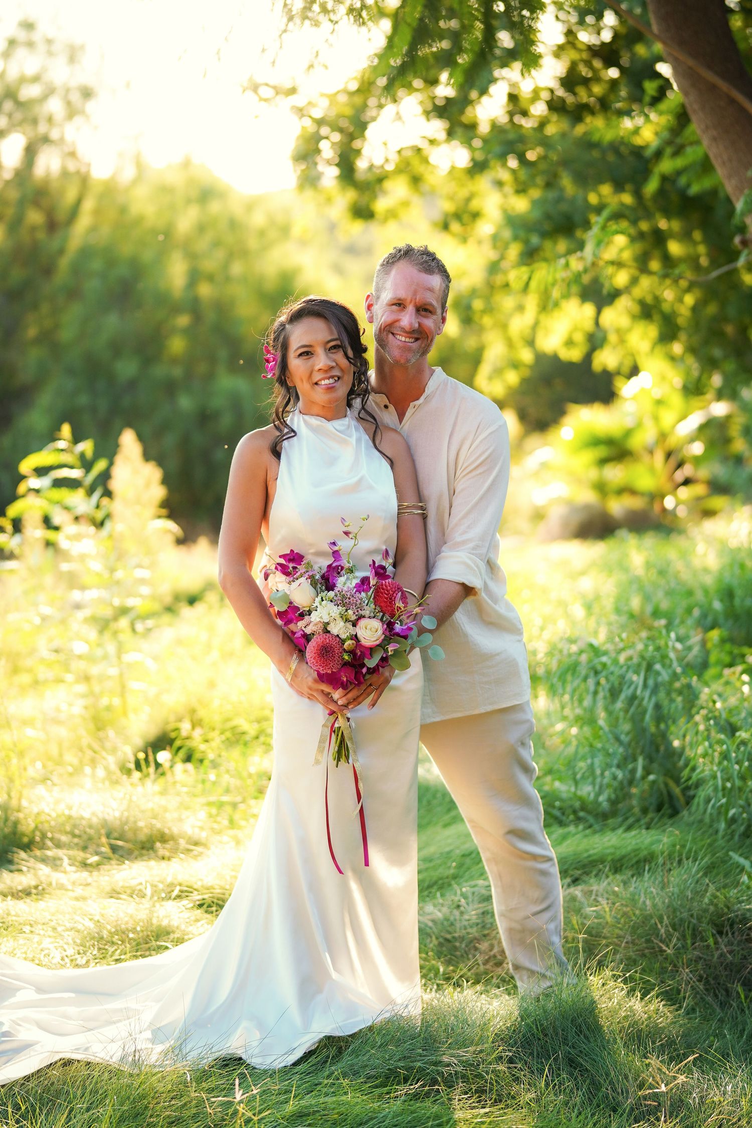Wedding couple embracing in a sunlit green meadow, the bride holding a vibrant pink and purple floral bouquet.