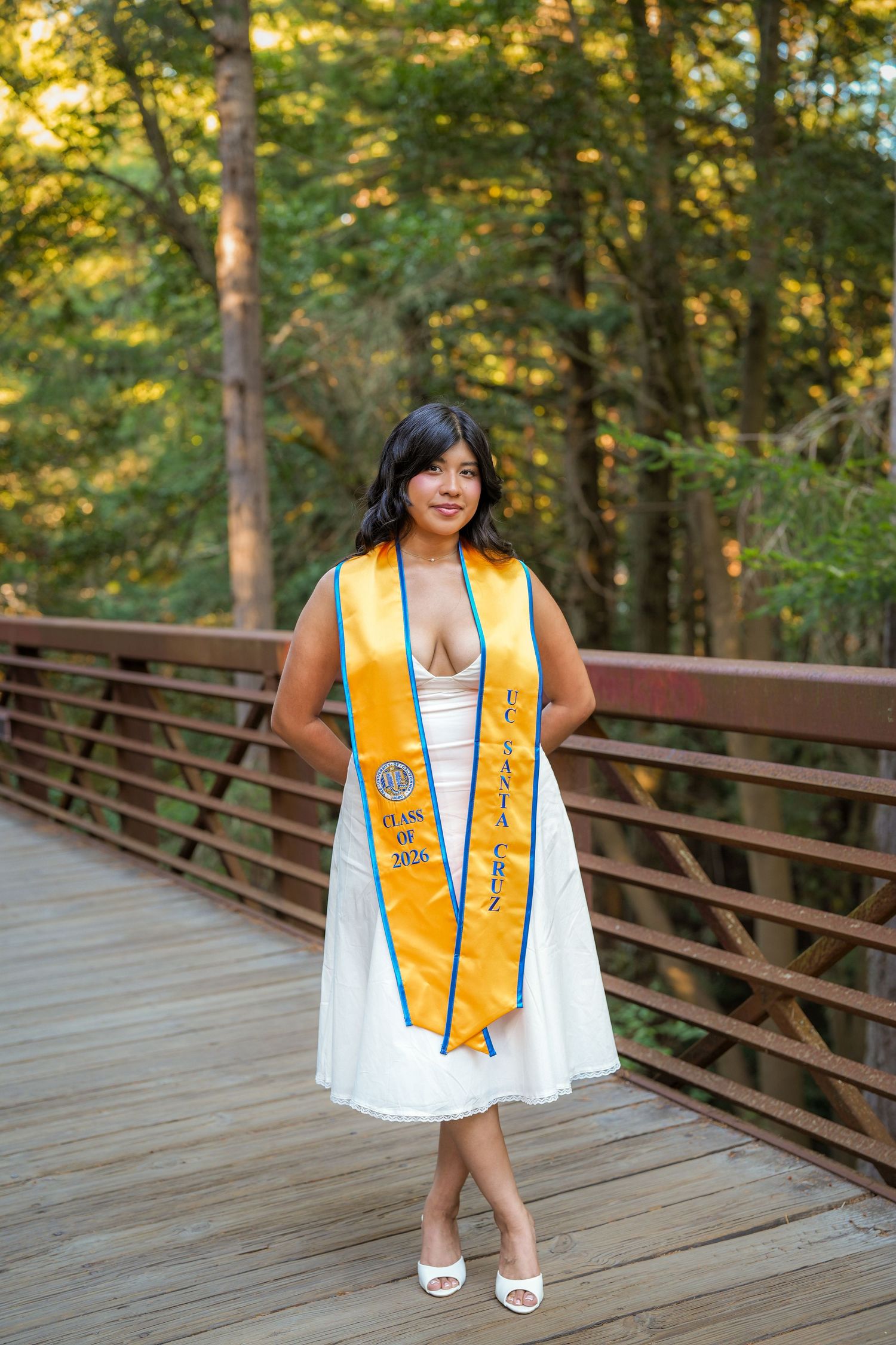 Graduate in a white dress and gold graduation sash stands smiling on a wooden bridge surrounded by sunlit trees.