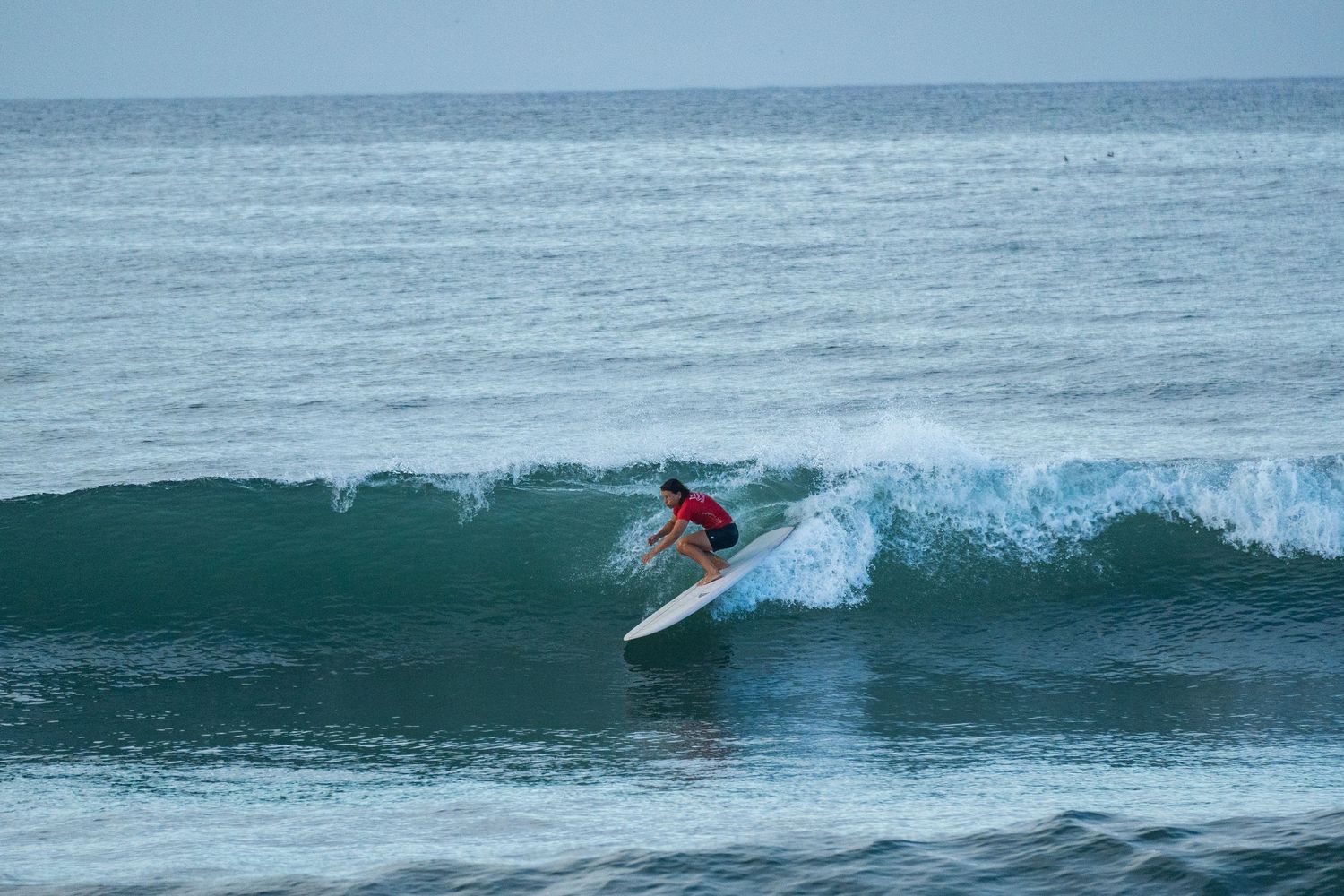 Surfer in a red top crouches low on a white board while riding a green wave in open ocean water.