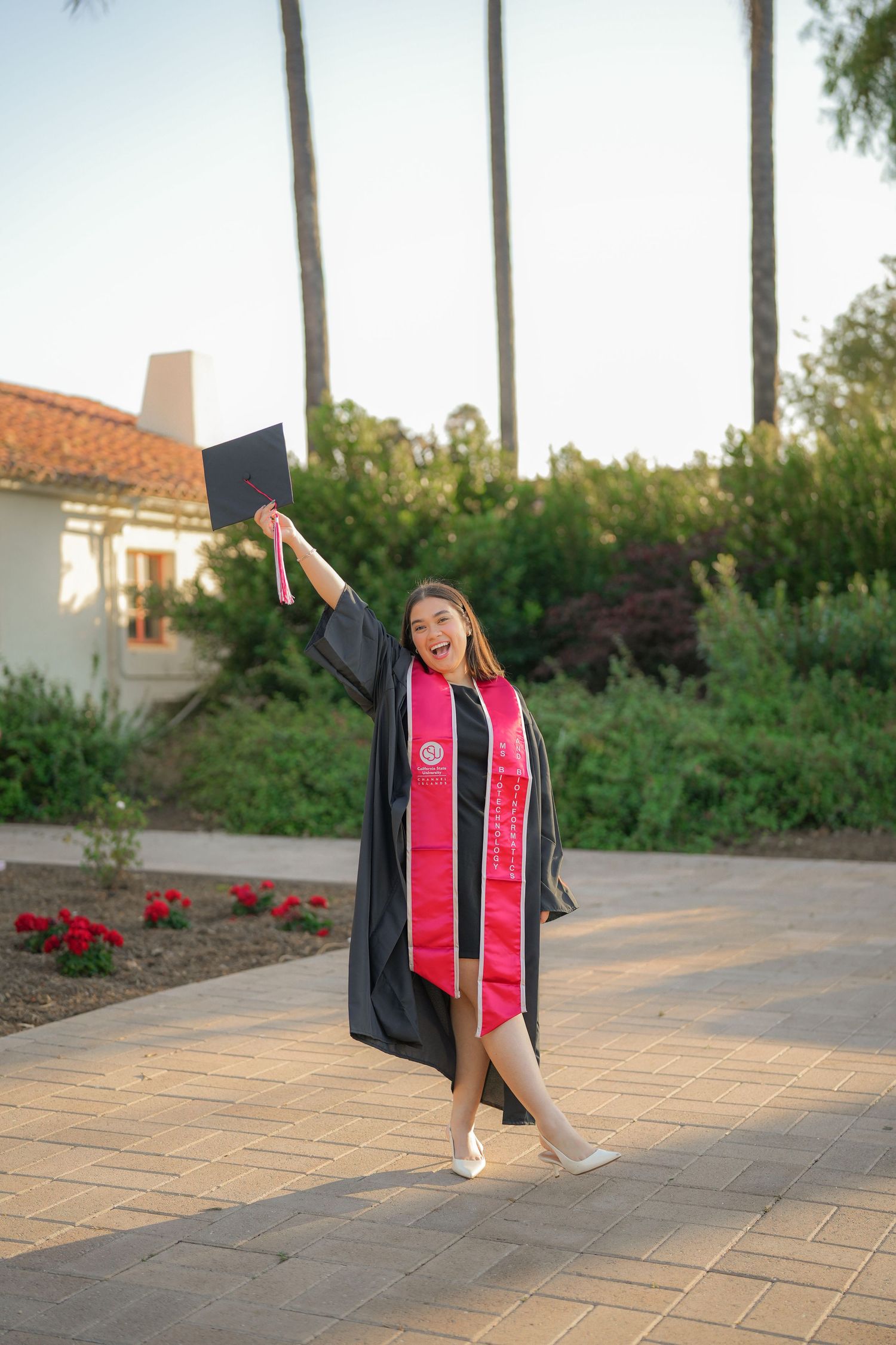 A joyful graduate in a black gown and red stole raises her graduation cap in the air on a brick path near blooming red flowers.