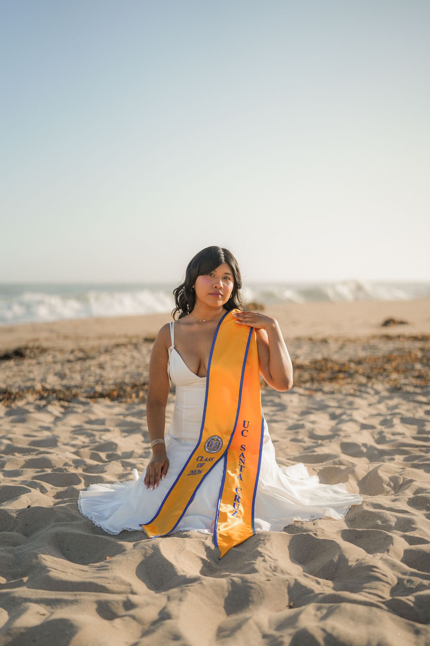 Graduate in a white dress with an orange UC Santa Cruz sash sitting on the sand with ocean waves behind her.