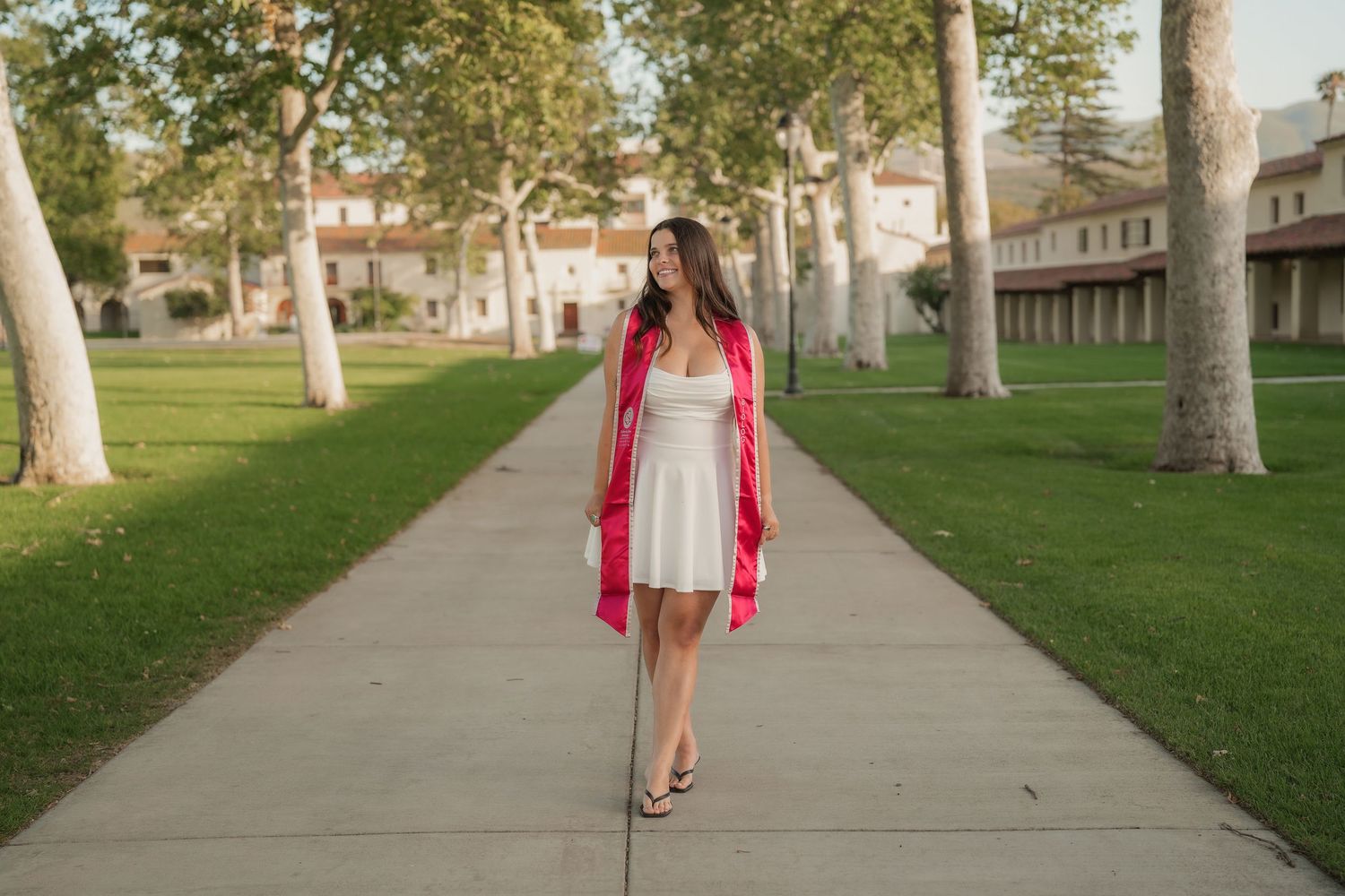 A graduate in a white dress and red stole walks down a sunny tree-lined campus path with grass and historic buildings behind her.
