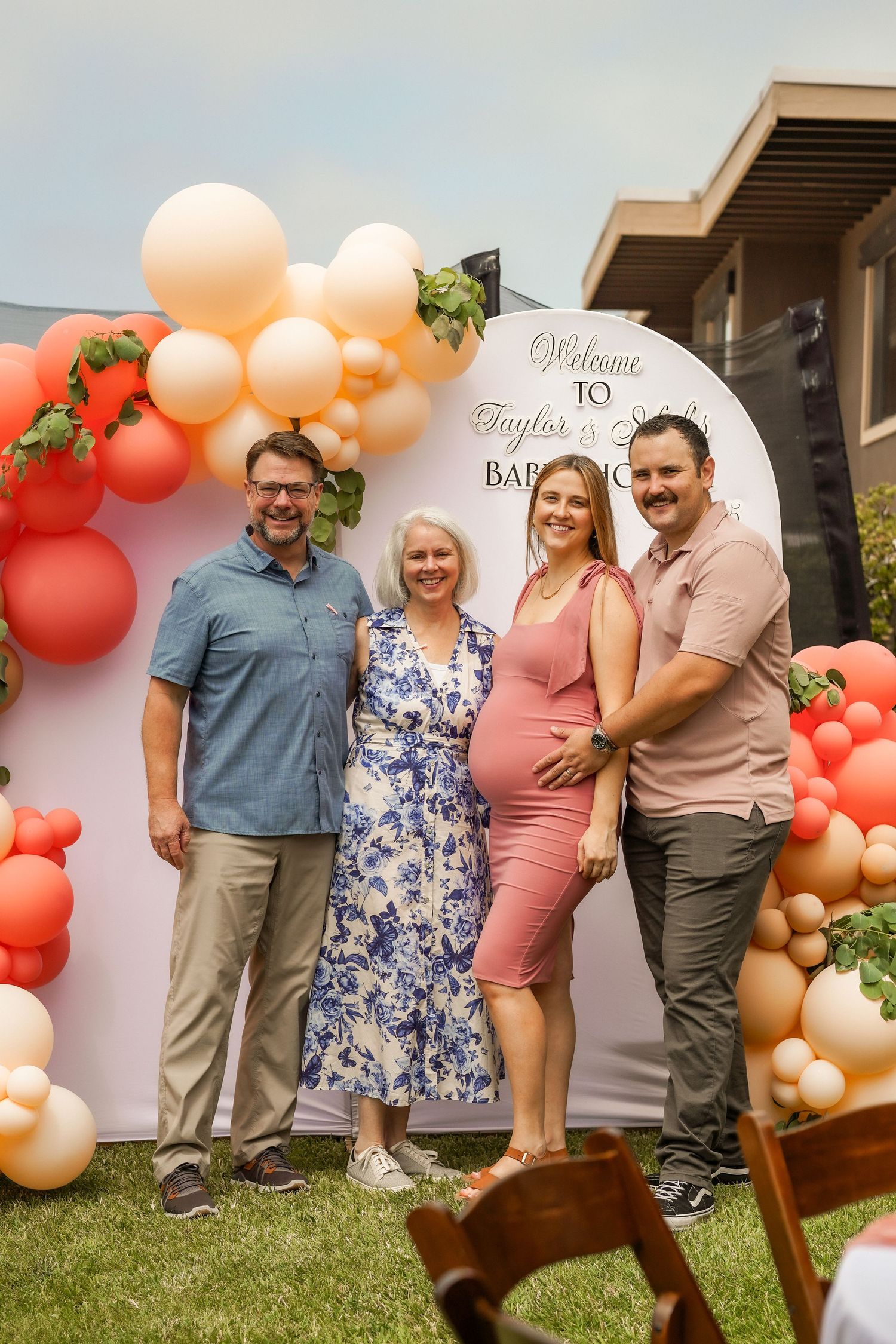 Four adults pose together outdoors beside a peach and coral balloon garland at a baby shower, with a pregnant woman in a pink dress.