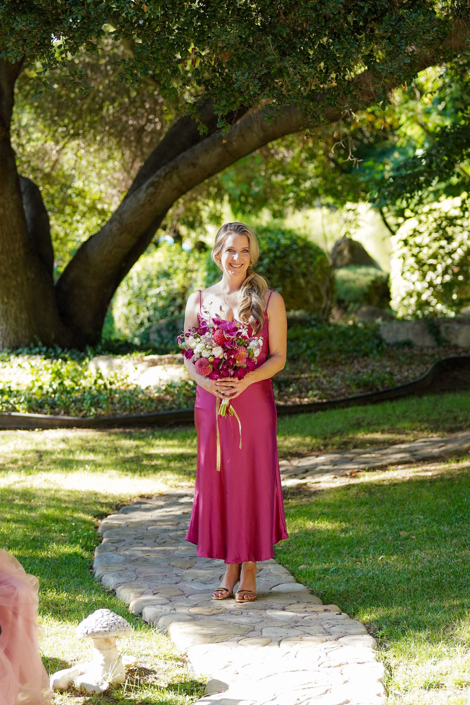 A bridesmaid in a magenta dress holds a pink bouquet while standing on a stone path beneath a large oak tree at a garden wedding.