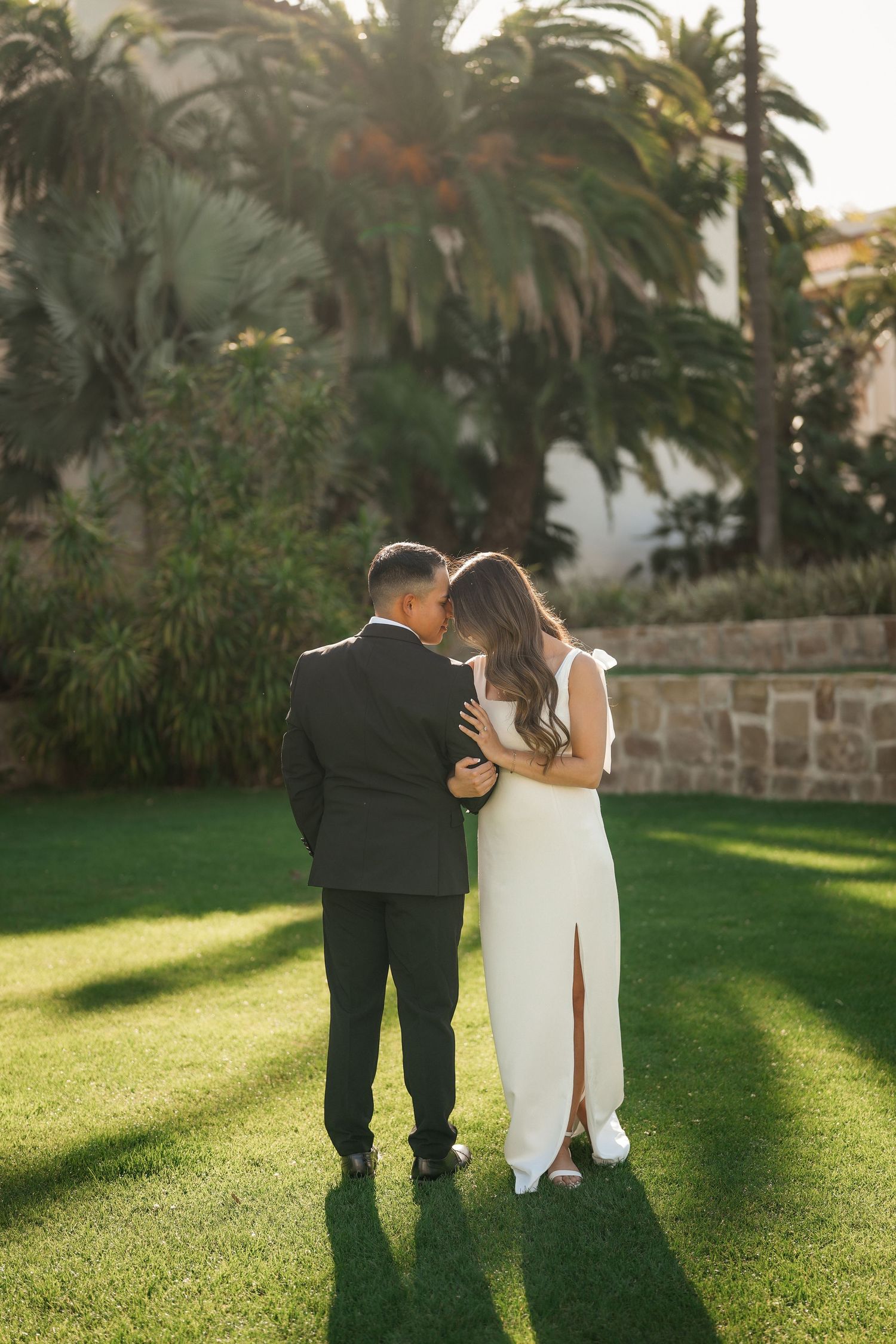 Bride and groom share a tender moment on a sunny lawn framed by palm trees and a stone garden wall.