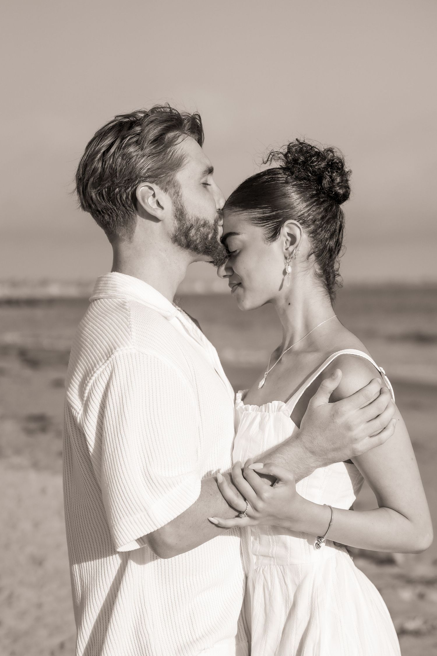 Sepia photo of a bearded man in a white shirt kissing his partner's forehead as they embrace on the beach.