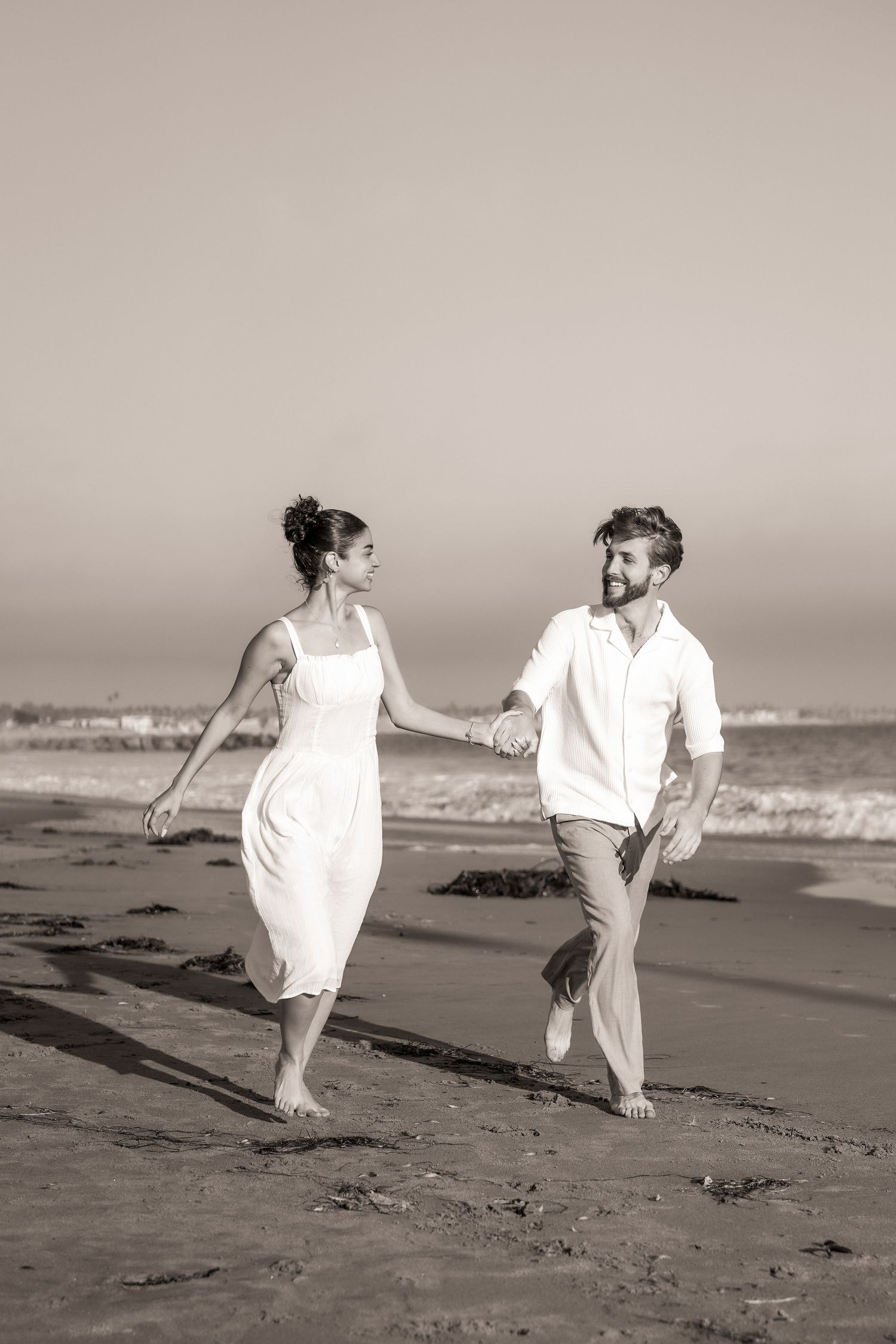 Sepia photo of a joyful couple in white outfits holding hands and running barefoot along the shoreline.